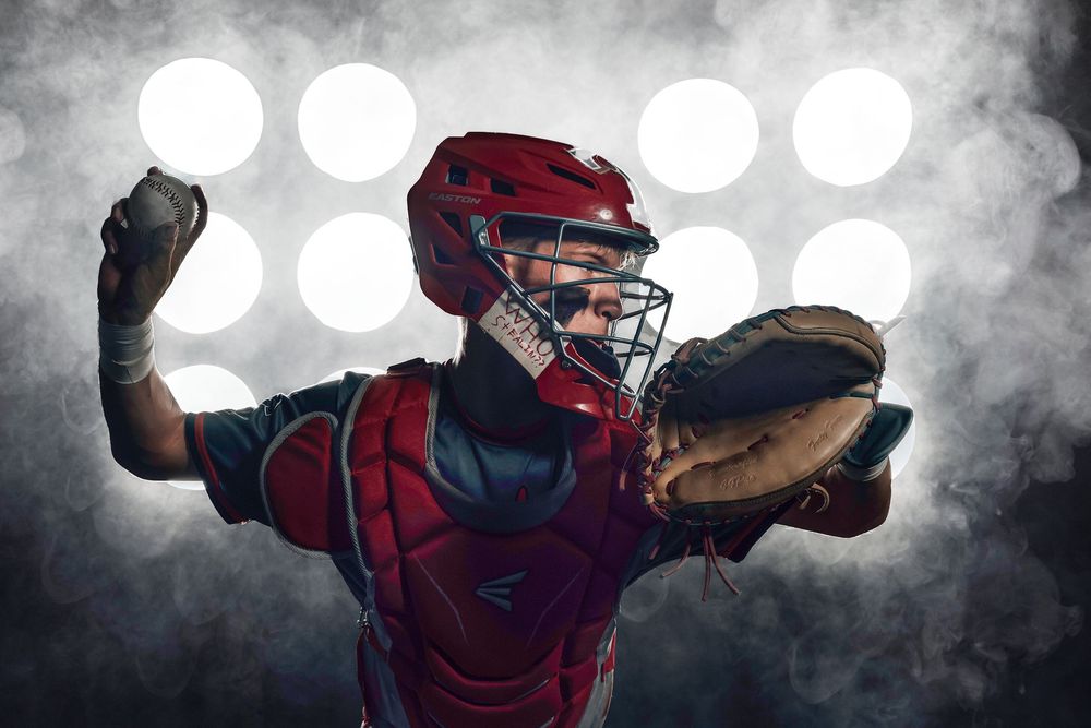 Baseball catcher in red protective gear against dramatic backlit background with stadium lights.