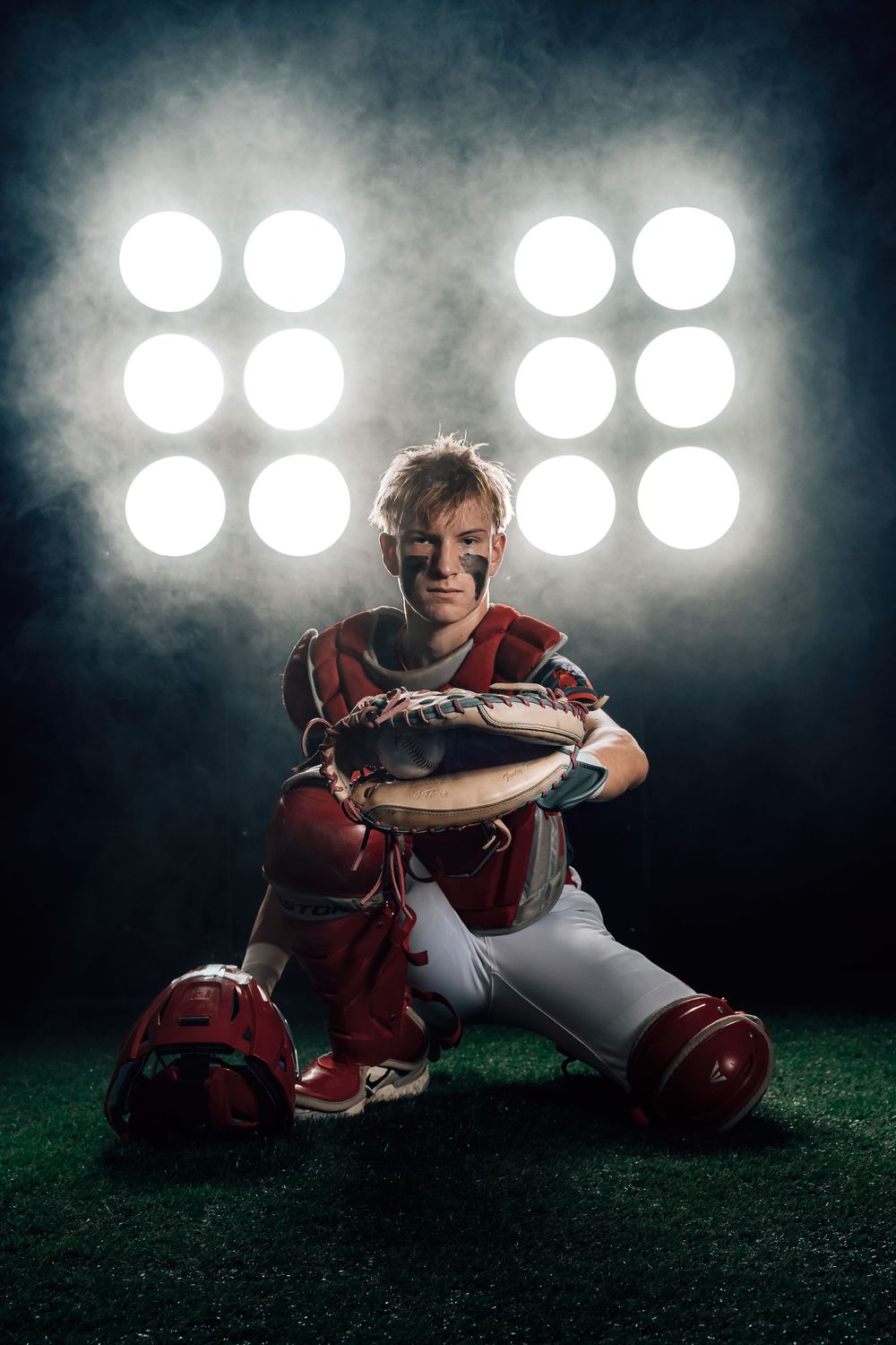 Baseball catcher in red uniform sits on field turf at night with stadium lights glowing dramatically in background.