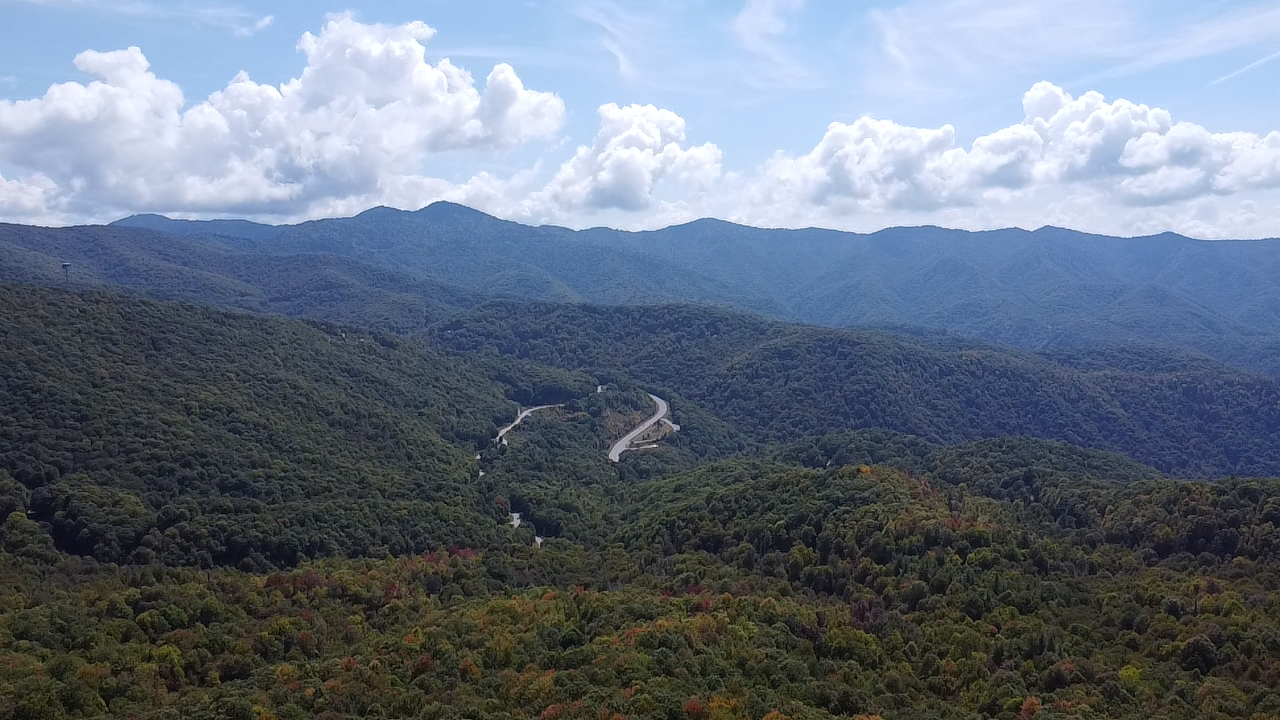 A panoramic view of winding mountain roads through forested peaks with blue skies and puffy white clouds overhead.