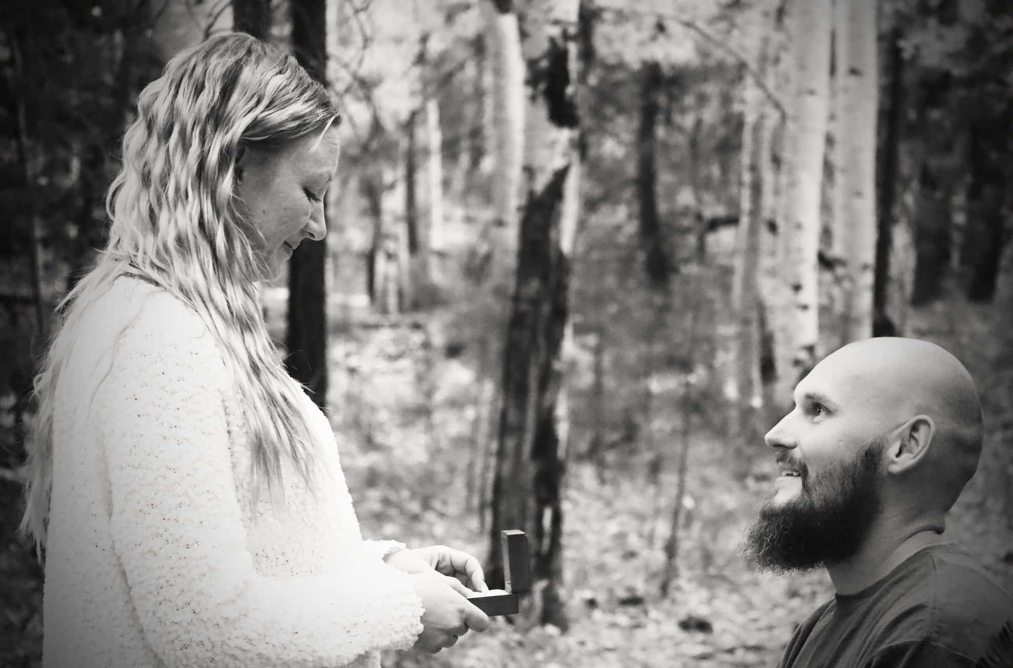 Black and white photo of a man on one knee proposing to a woman in a forest.