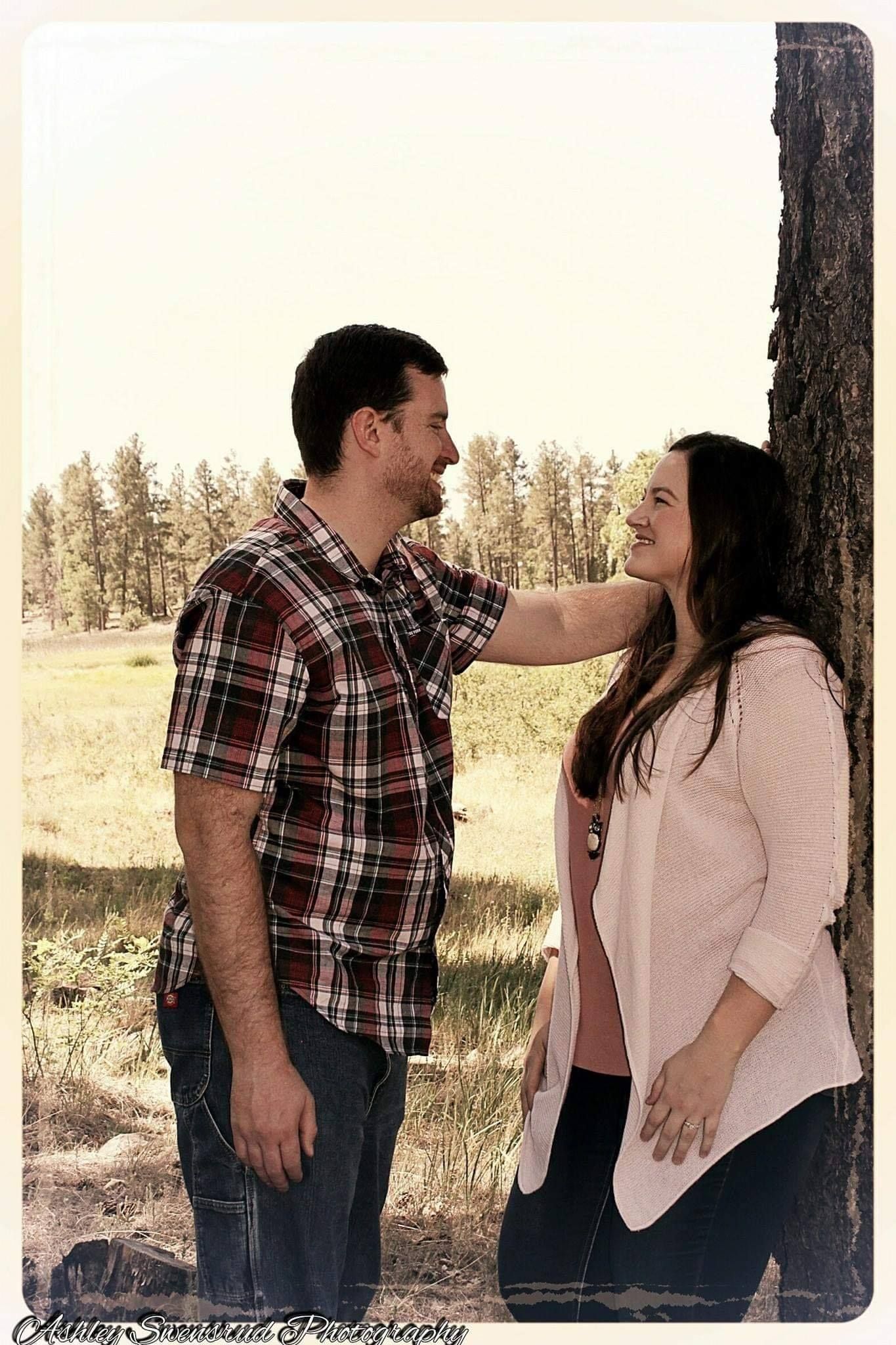 A smiling couple shares a tender moment leaning against a tree in a sunlit meadow.