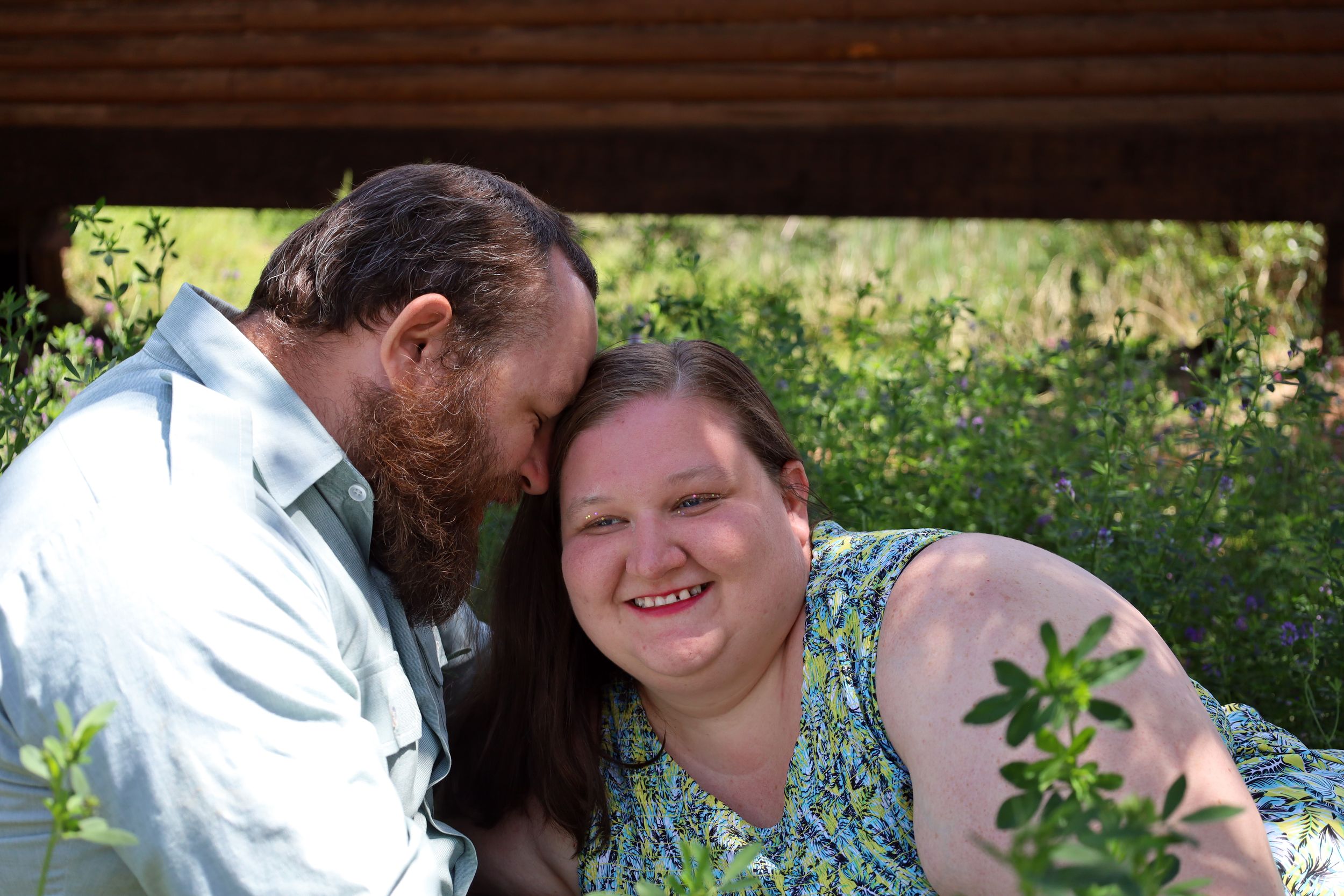 A bearded man kisses a smiling woman on the cheek while sitting outdoors among green plants.