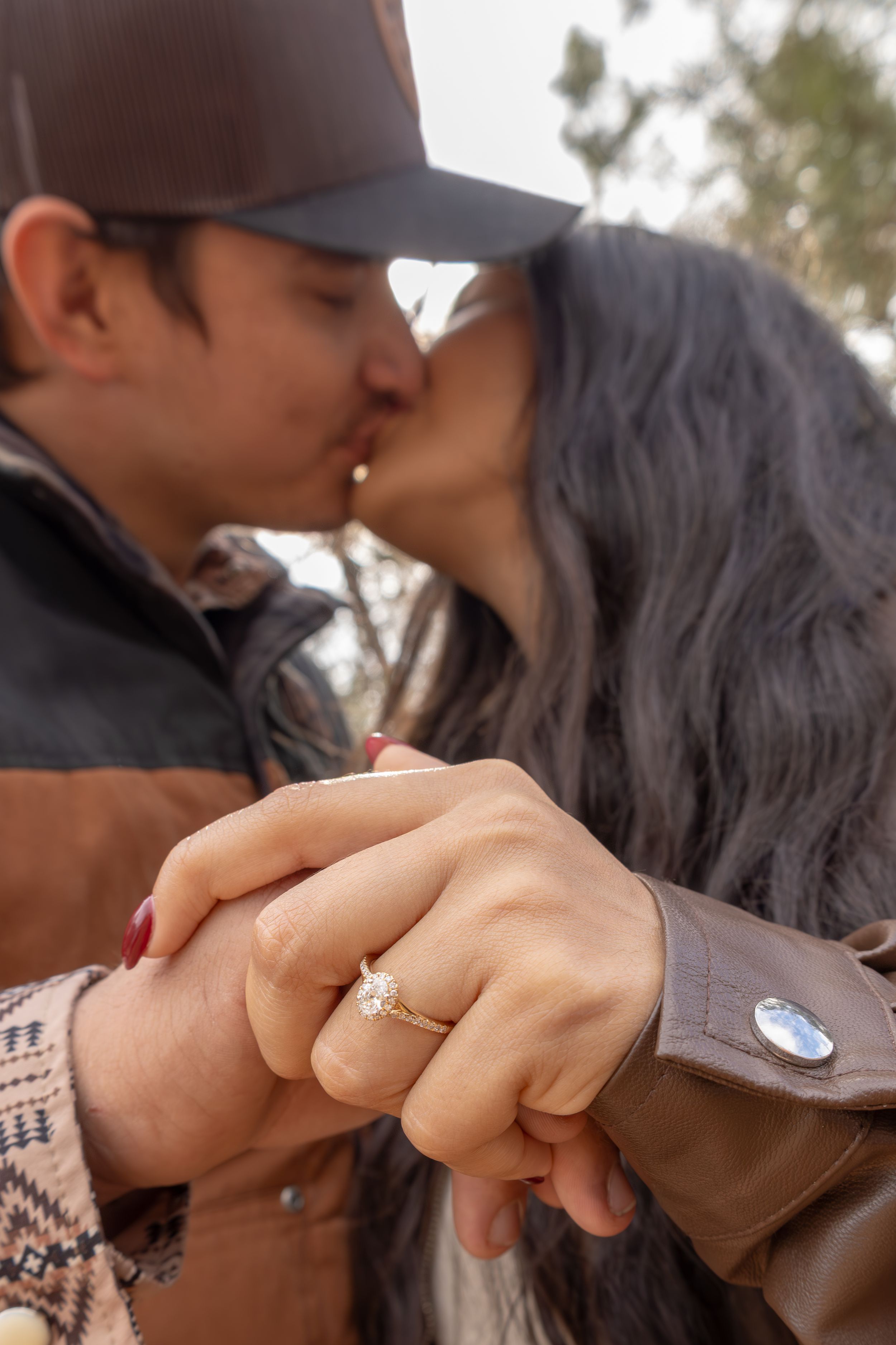 Engaged couple kissing while showcasing a diamond engagement ring in a forest setting.