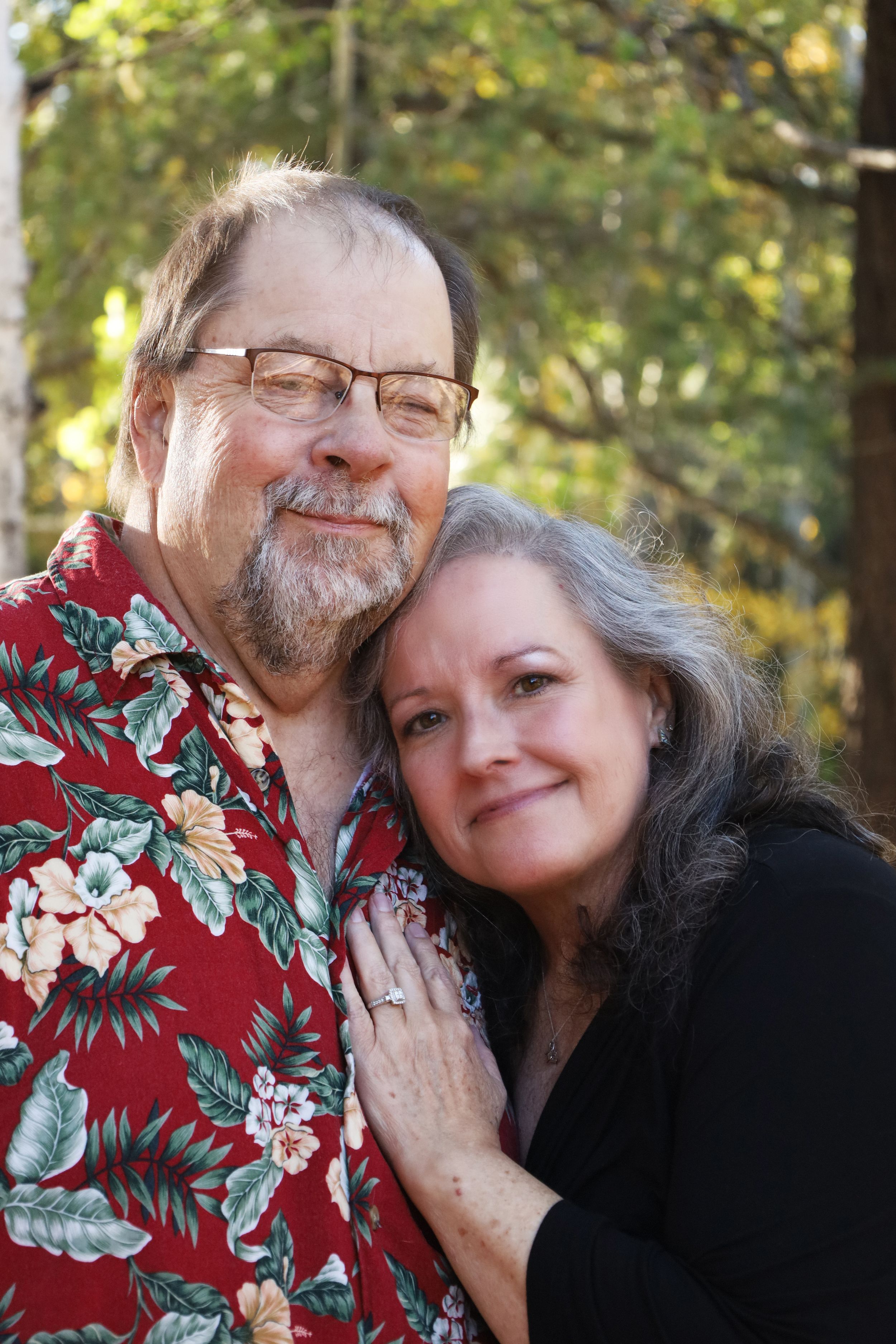 An older couple smiles warmly together outdoors, the woman wearing a diamond ring on her hand.