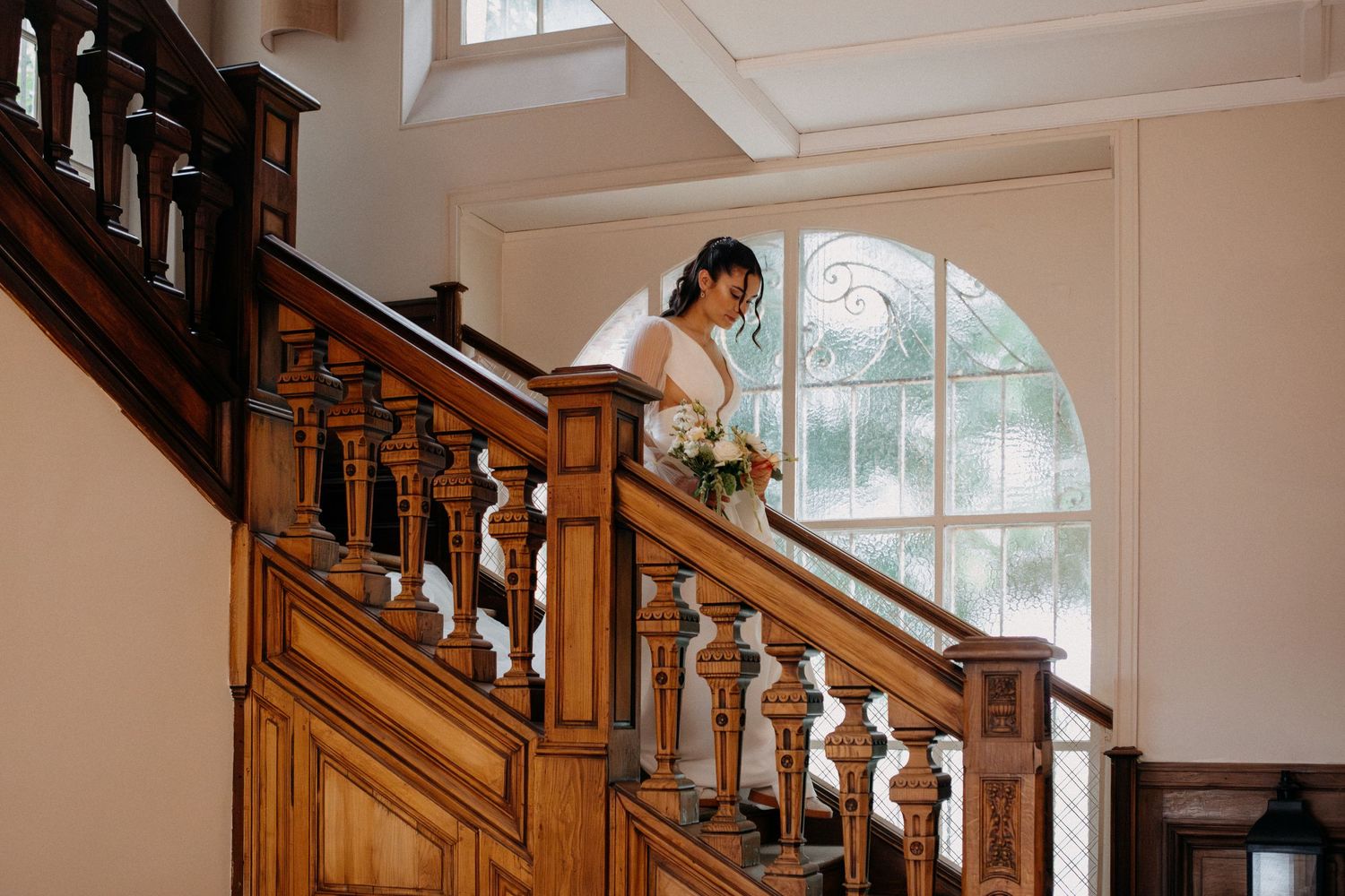 Bride in white gown holding a bouquet while descending an elegant wooden staircase.