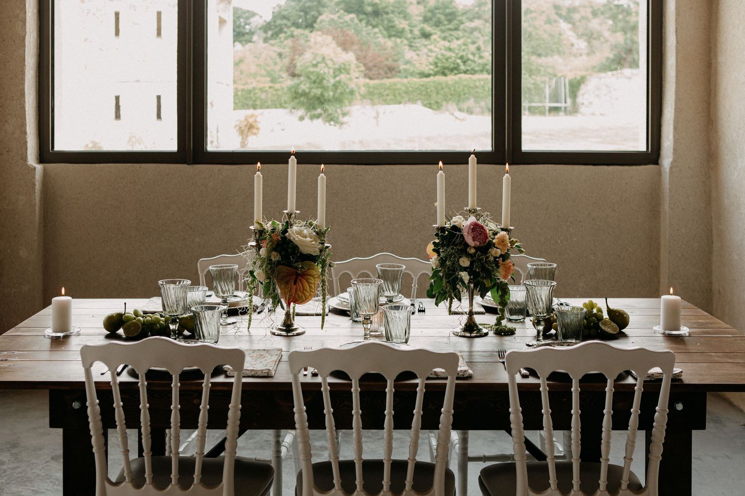 Rustic wedding reception table with taper candles and floral centerpieces by large windows.