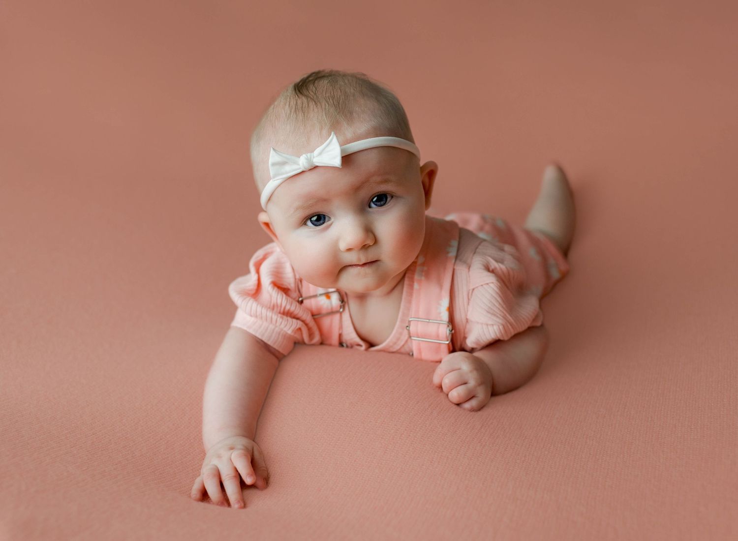 Baby wearing white headband and pink outfit lying on peach-colored backdrop during professional photo session.