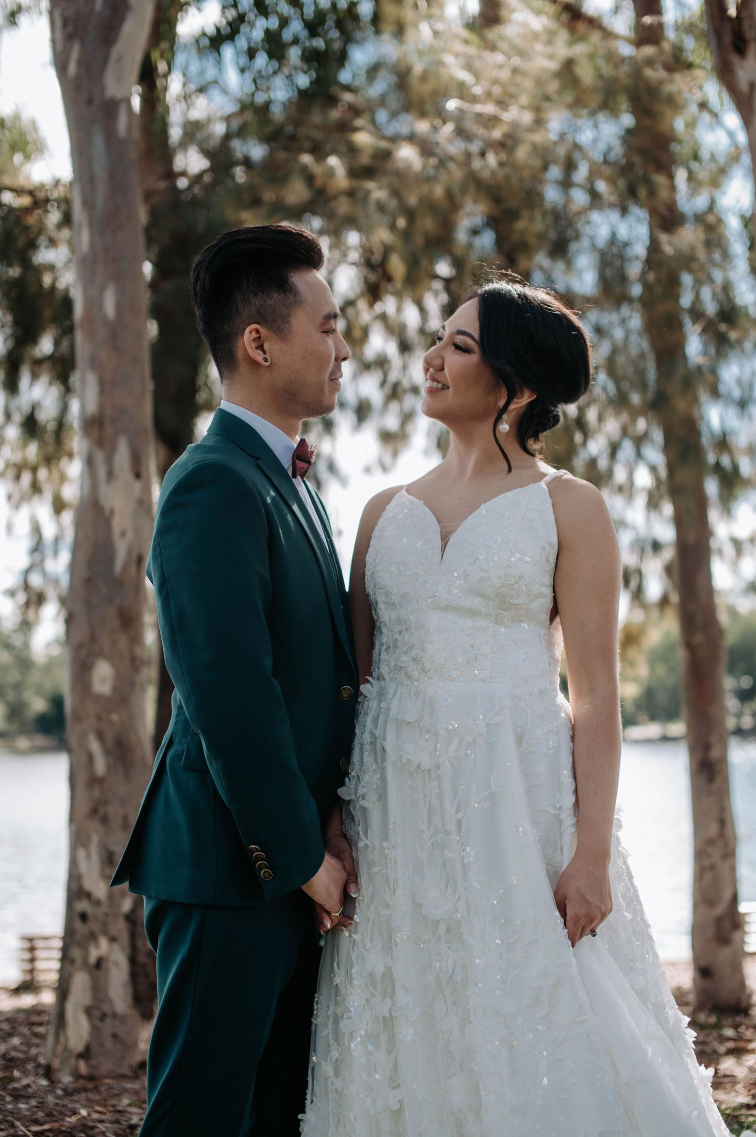 Romantic newlywed couple in white wedding dress and dark teal suit pose among trees in an outdoor garden setting.