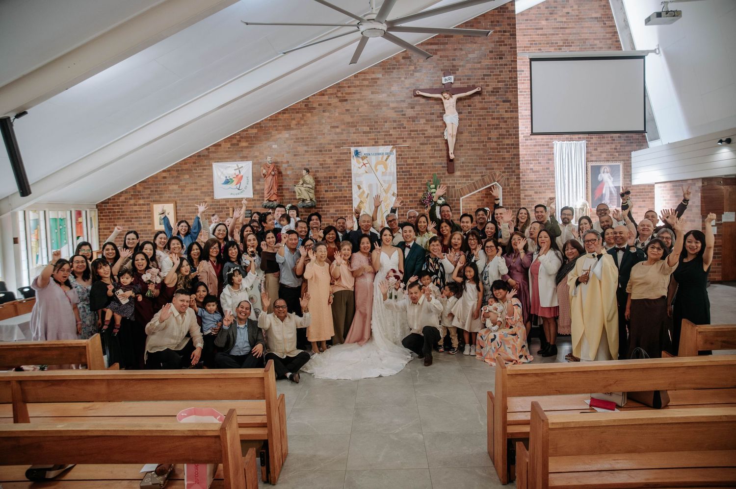 Large wedding ceremony group photo showing guests standing together in a modern church with brick walls and wooden pews.