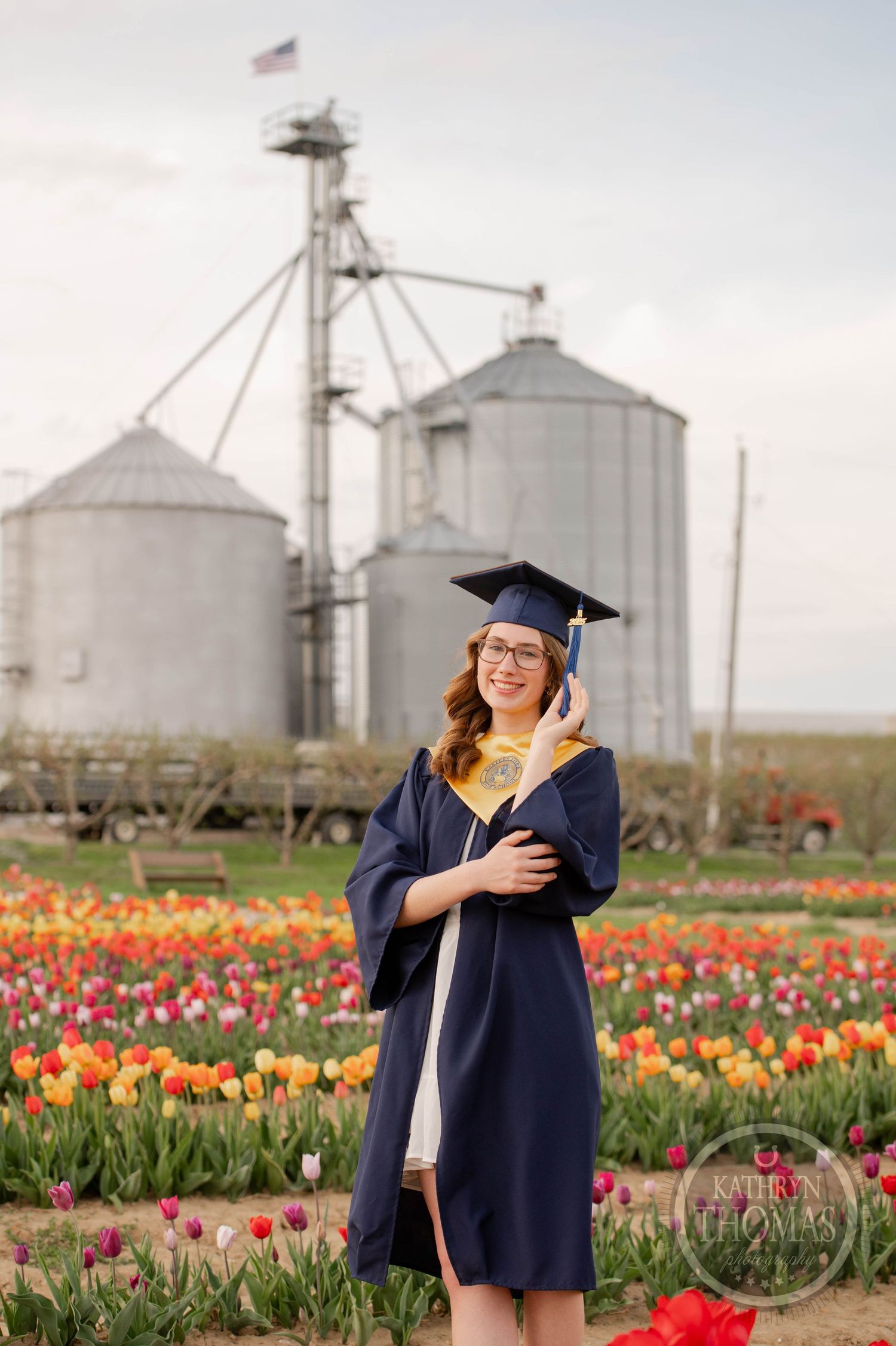 Graduate in navy cap and gown poses in field of colorful tulips with grain silos in background.