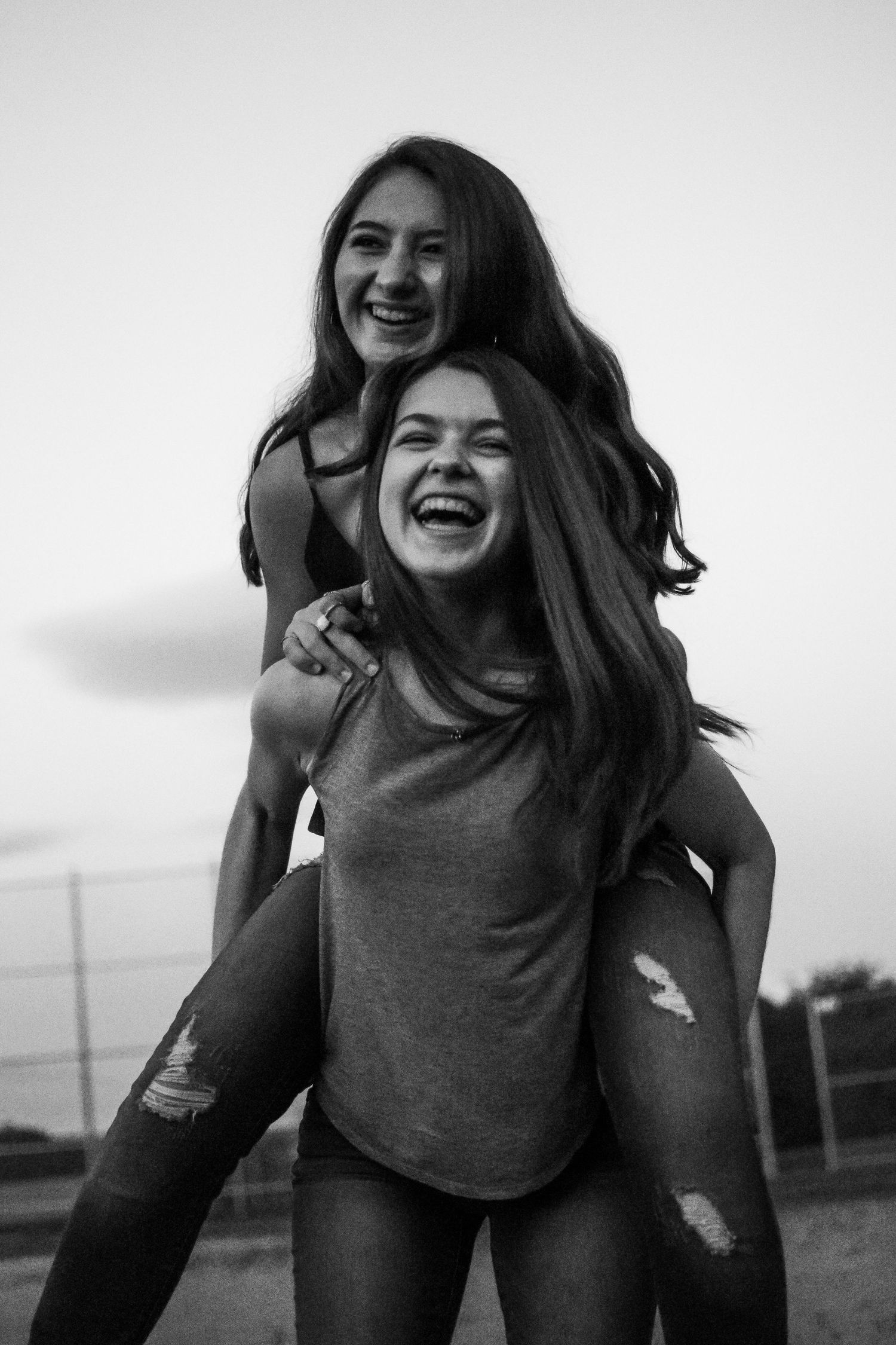 A tender black and white photograph of two smiling friends. One girl is getting a piggy back ride from her friend.