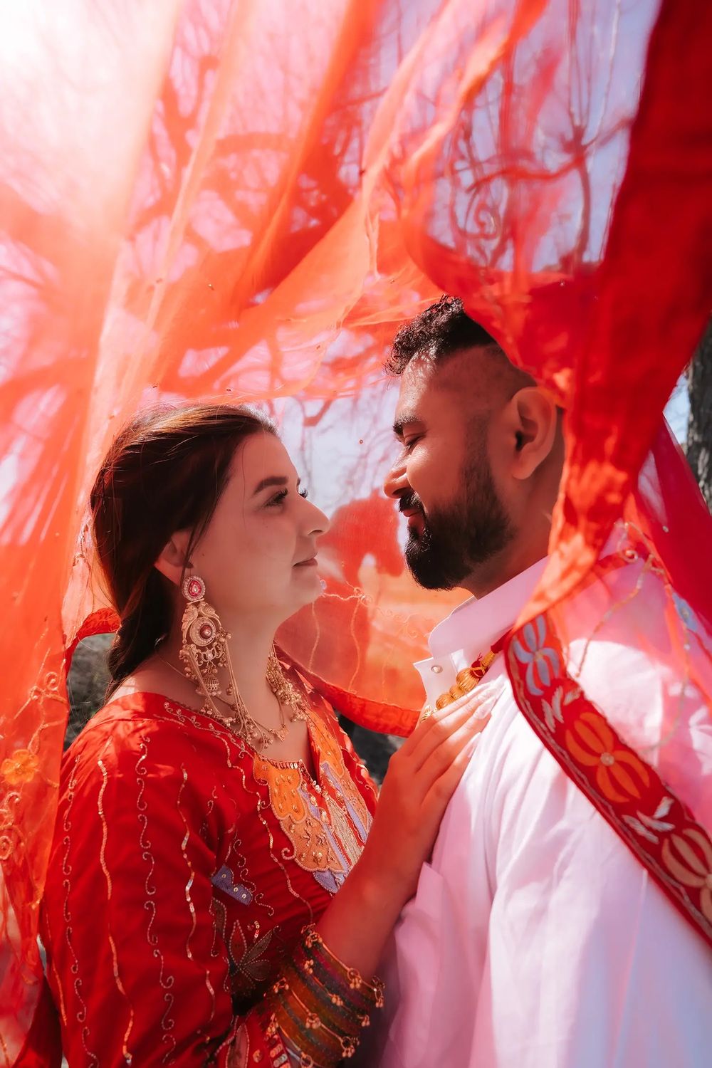 A couple in traditional Indian wedding attire gazes lovingly at each other beneath a flowing red dupatta veil.