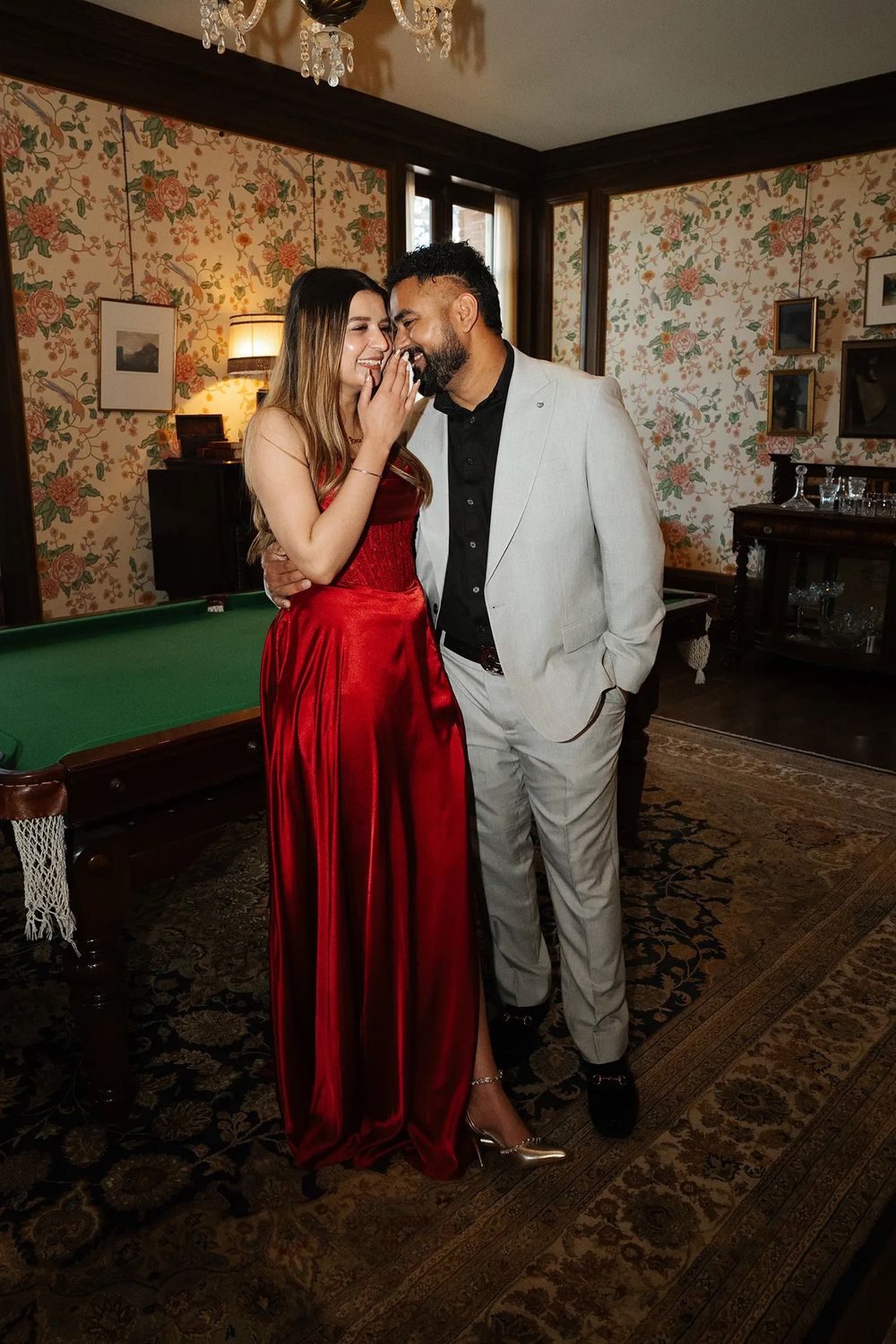 Couple in elegant attire posing in a vintage room with floral wallpaper and a pool table.
