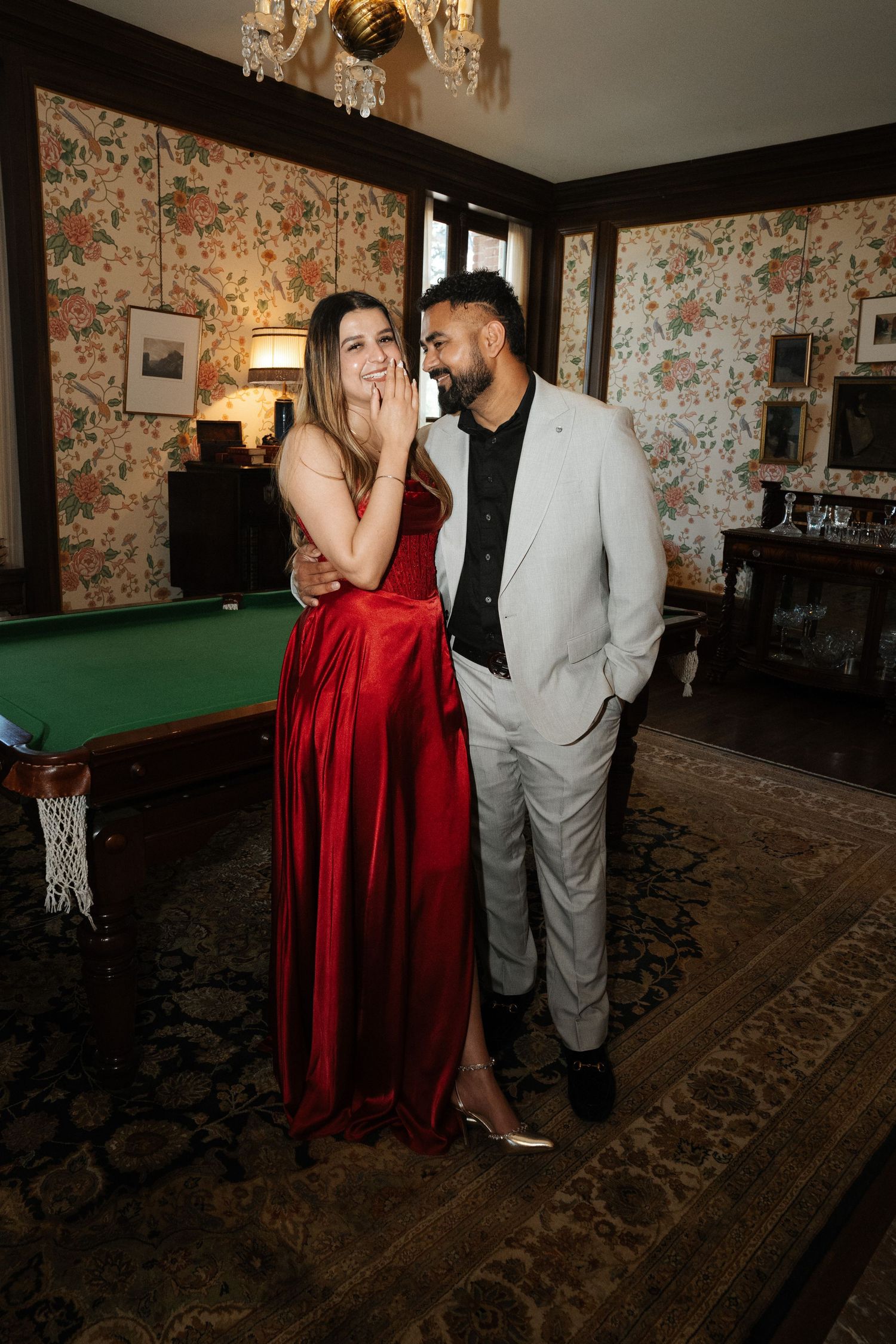 Couple in formal wear, woman in red gown and man in grey suit, posing in elegant room with pool table.