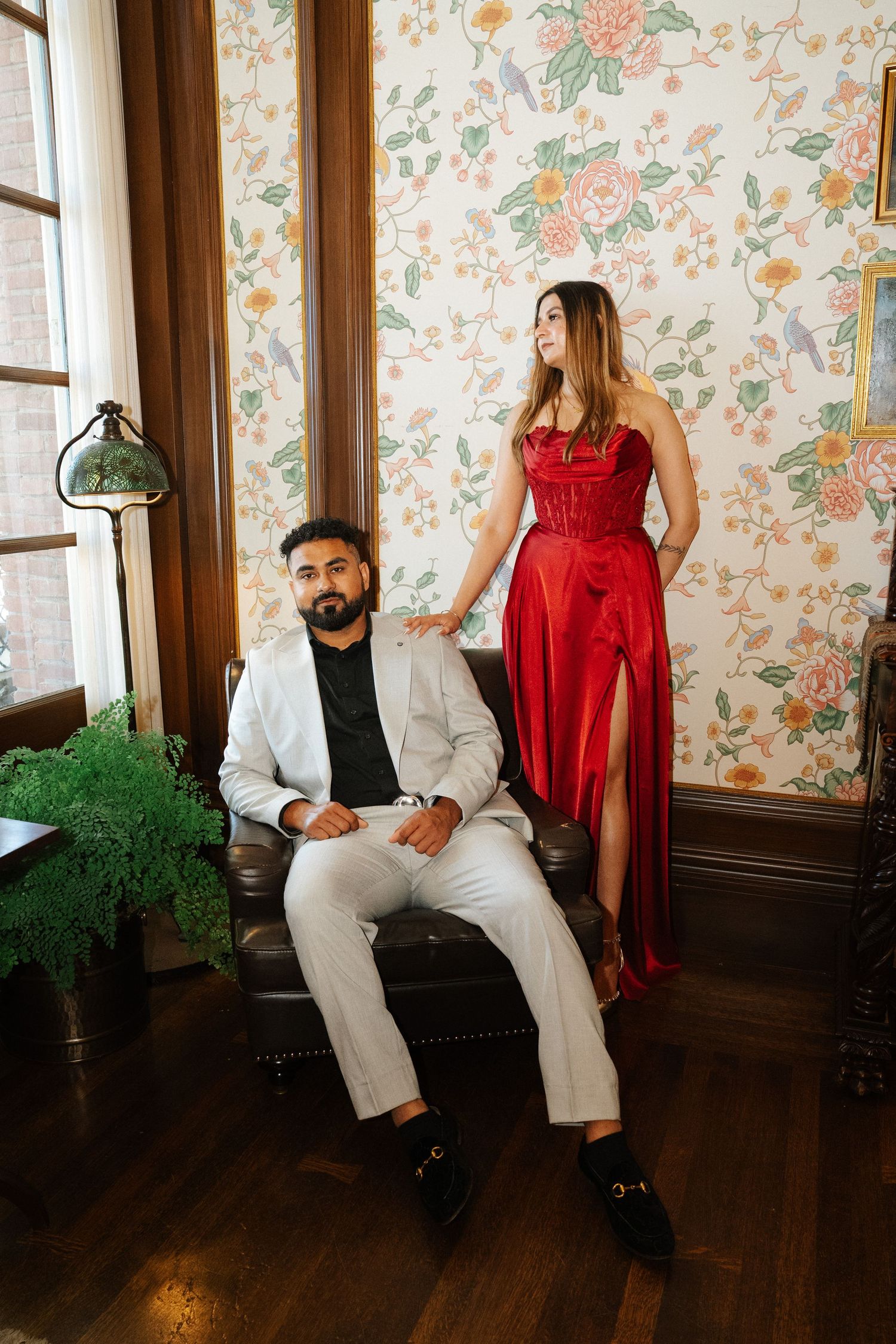 Man in white suit seated in leather chair beside woman in red satin gown, posing against floral wallpaper backdrop.