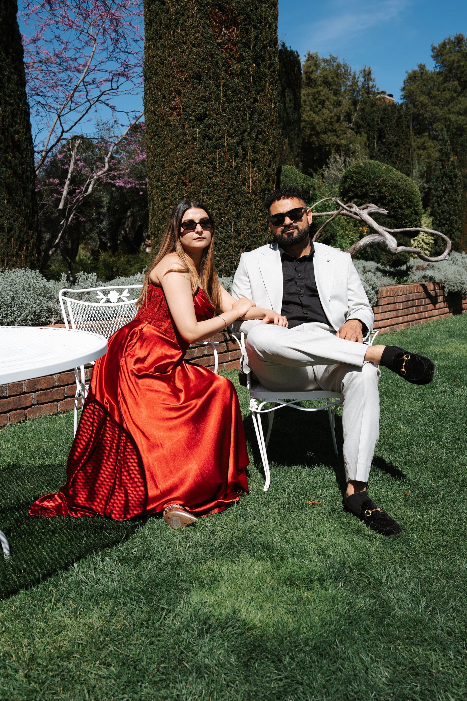 Stylish couple in sunglasses sitting in a sunny garden, woman in red satin gown, man in white suit.