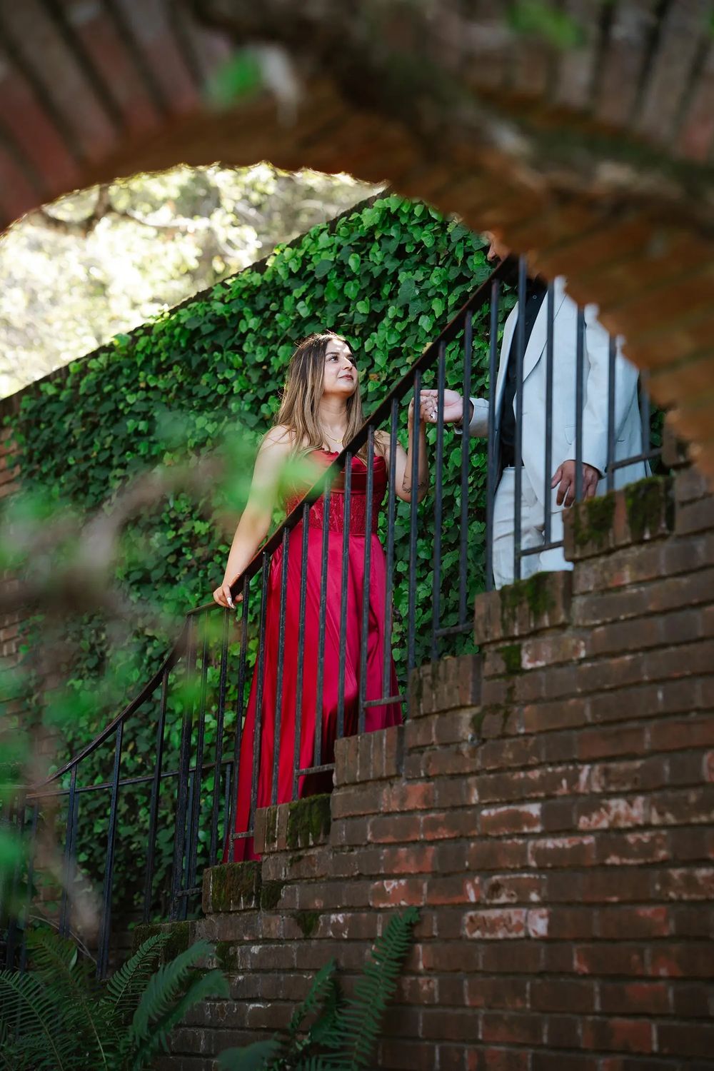 Woman in red striped dress standing on brick stairs under ivy-covered arch in lush garden setting.