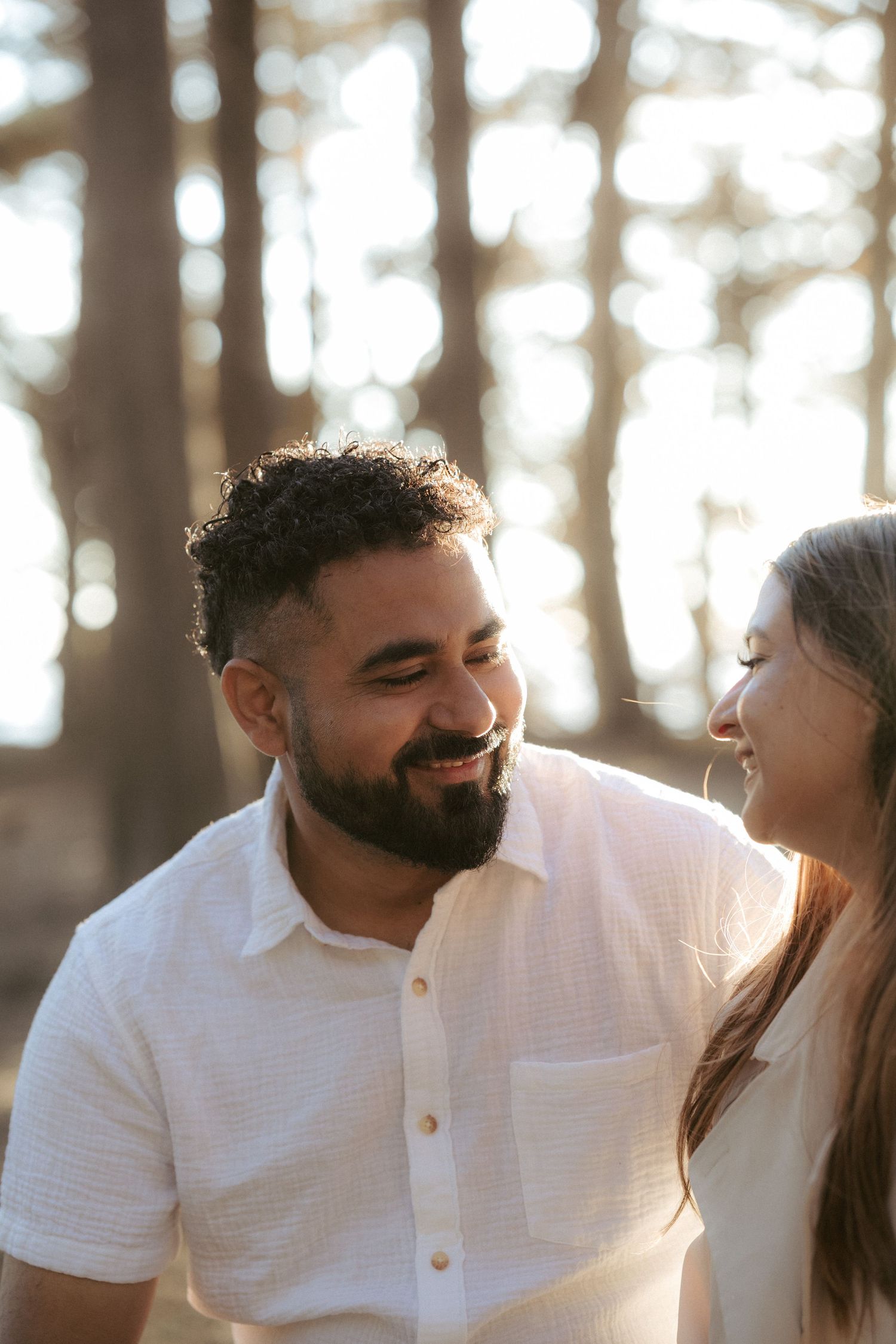 Smiling bearded man in white shirt sharing a joyful moment with a woman outdoors in soft light.