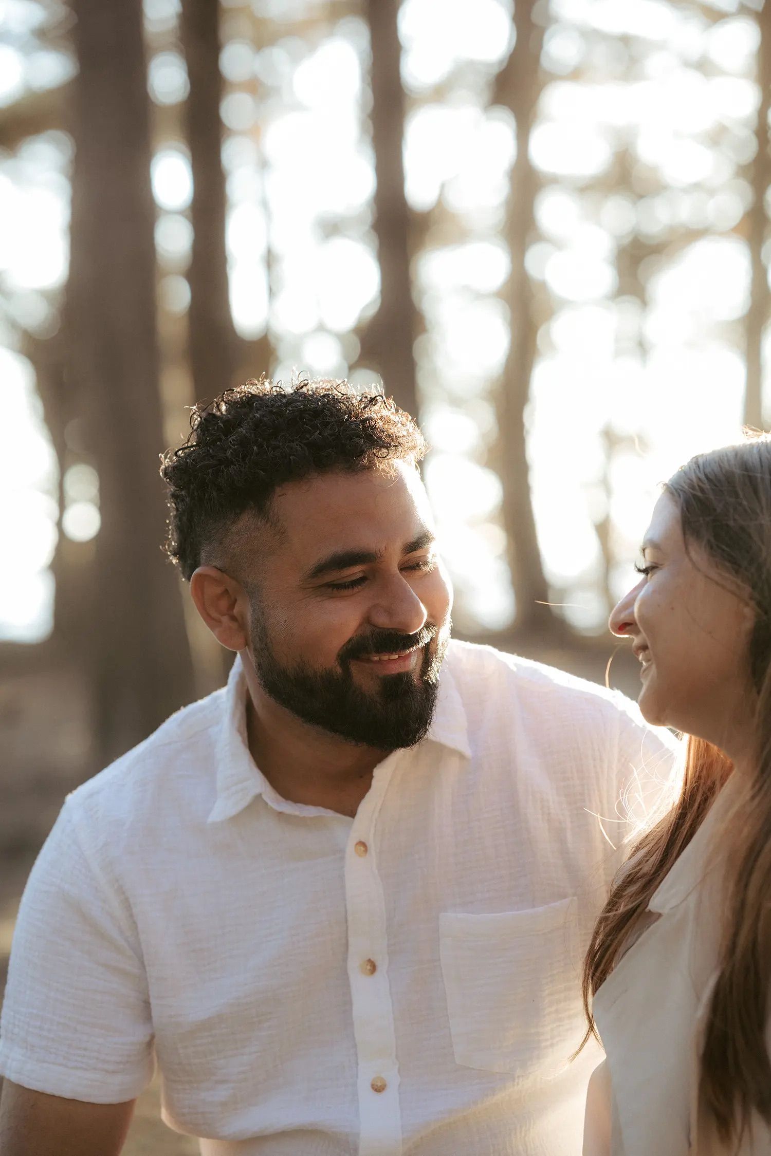 A smiling bearded man in a white shirt shares a joyful moment with a woman outdoors among tall trees.