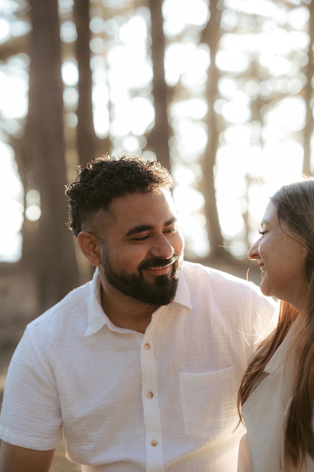 A smiling bearded man in a white shirt shares a joyful moment with a woman outdoors among tall trees.