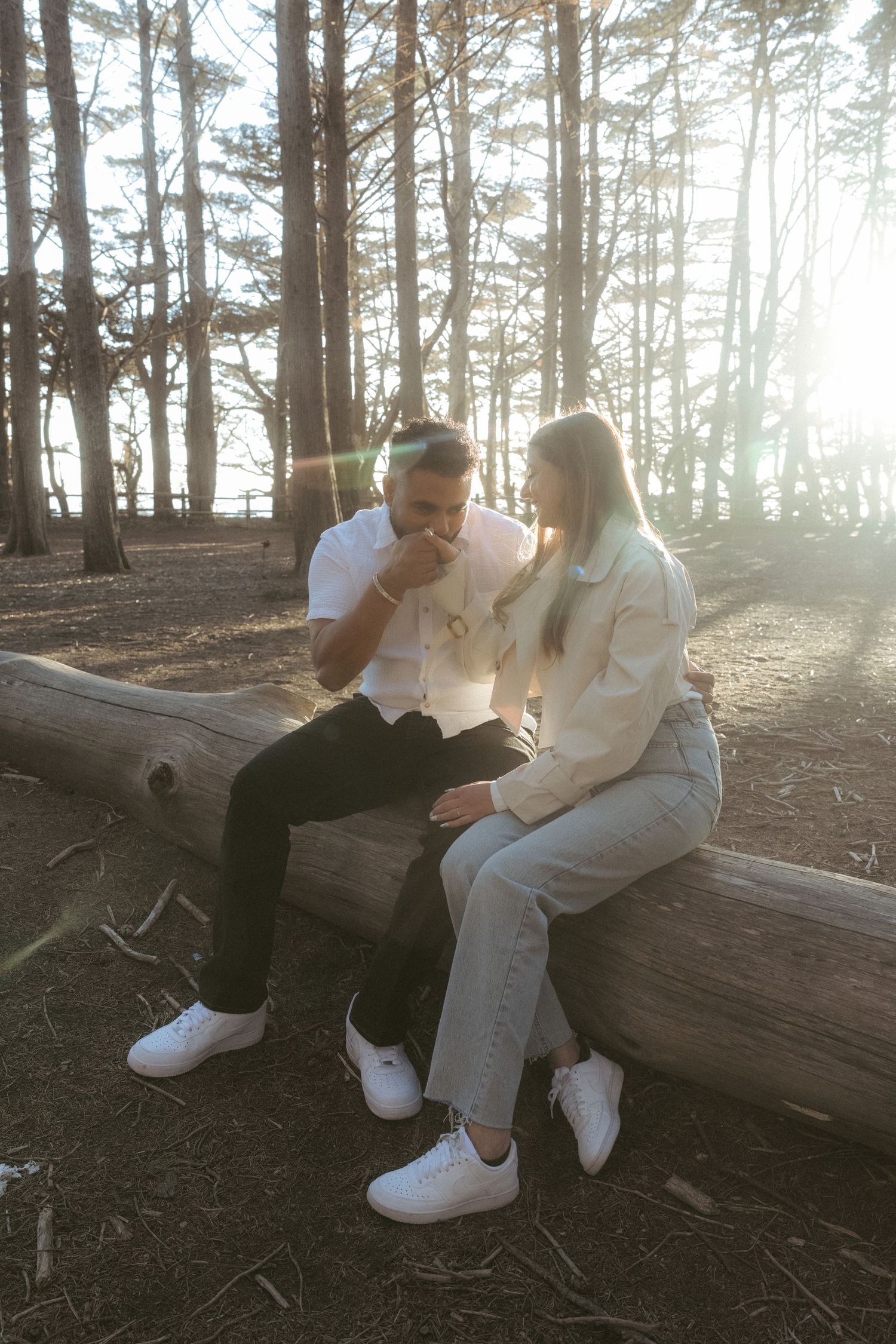 Couple sitting on a fallen log in a sunlit forest, wearing white tops and jeans with white sneakers.