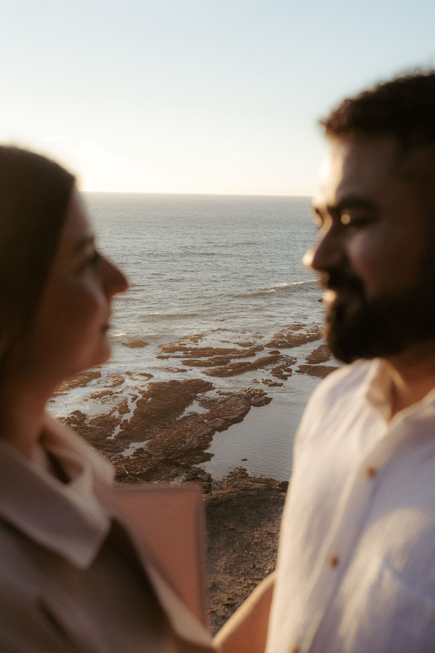 Couple sharing a romantic moment by the ocean at sunset, with rocky coastline and calm sea in the background.