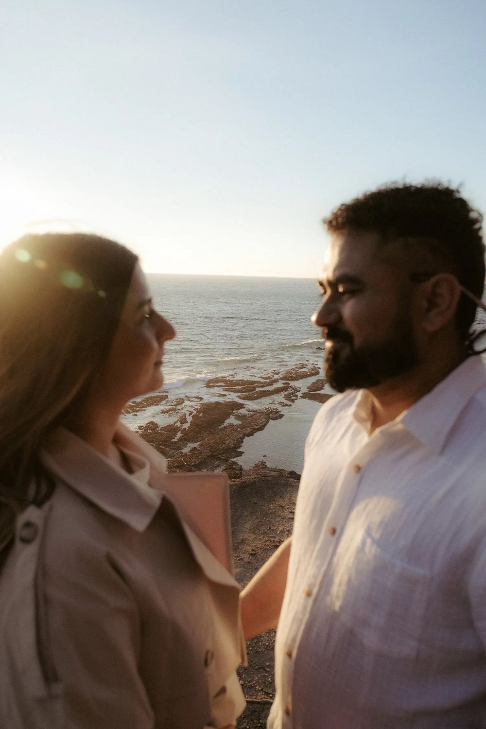 Couple sharing a romantic moment by the ocean at sunset, with golden light and rocky coastline in the background.