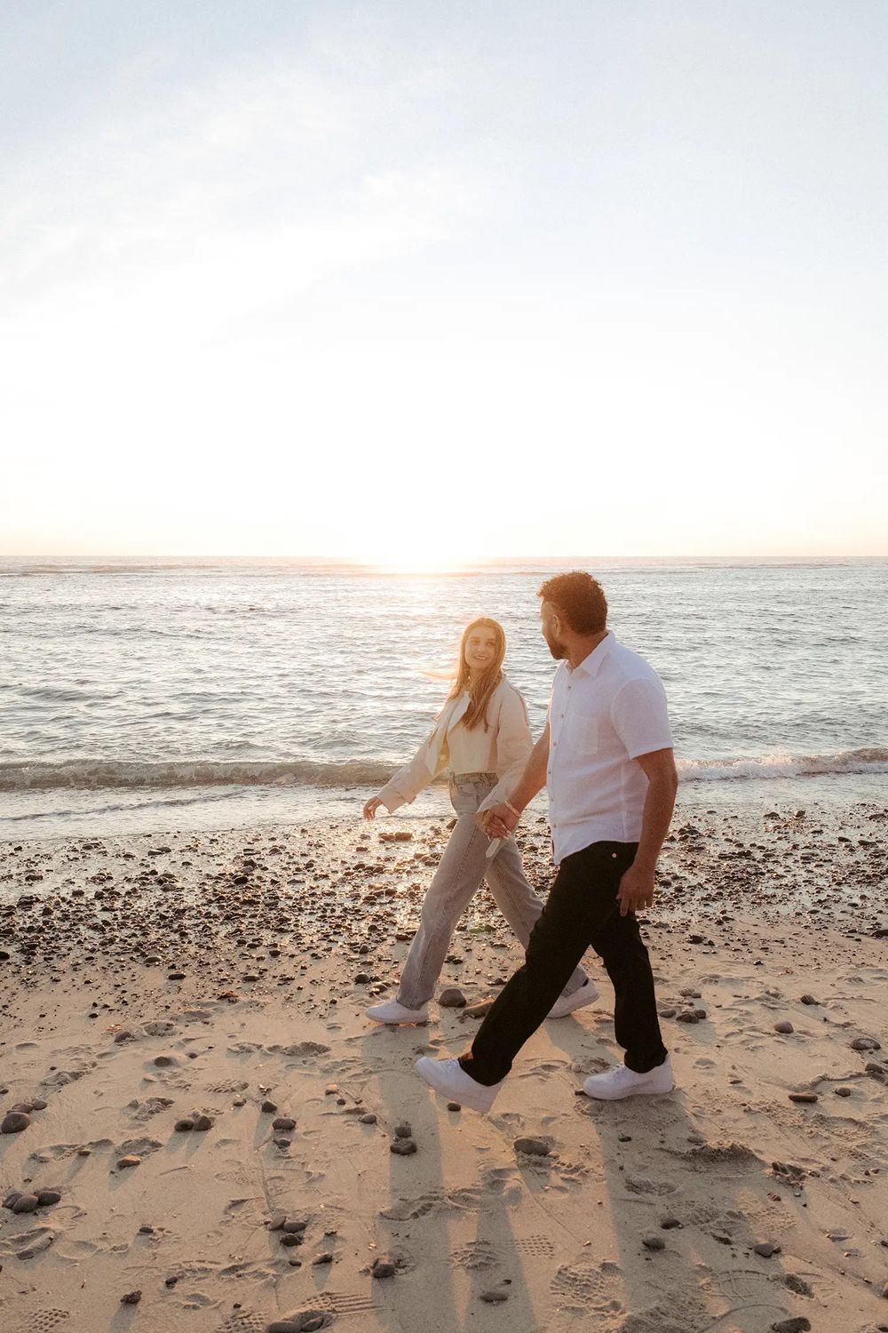 Couple walking hand in hand on a sandy beach at sunset, wearing casual white tops and dark pants.