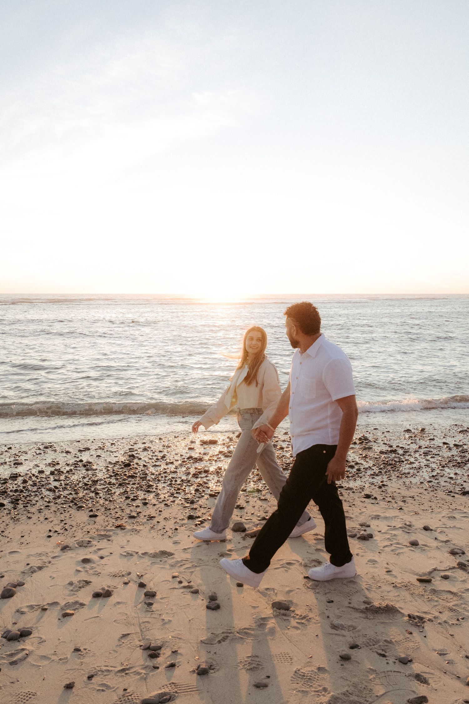 Couple walking hand in hand on a sandy beach at sunset, with gentle waves and golden light in the background.