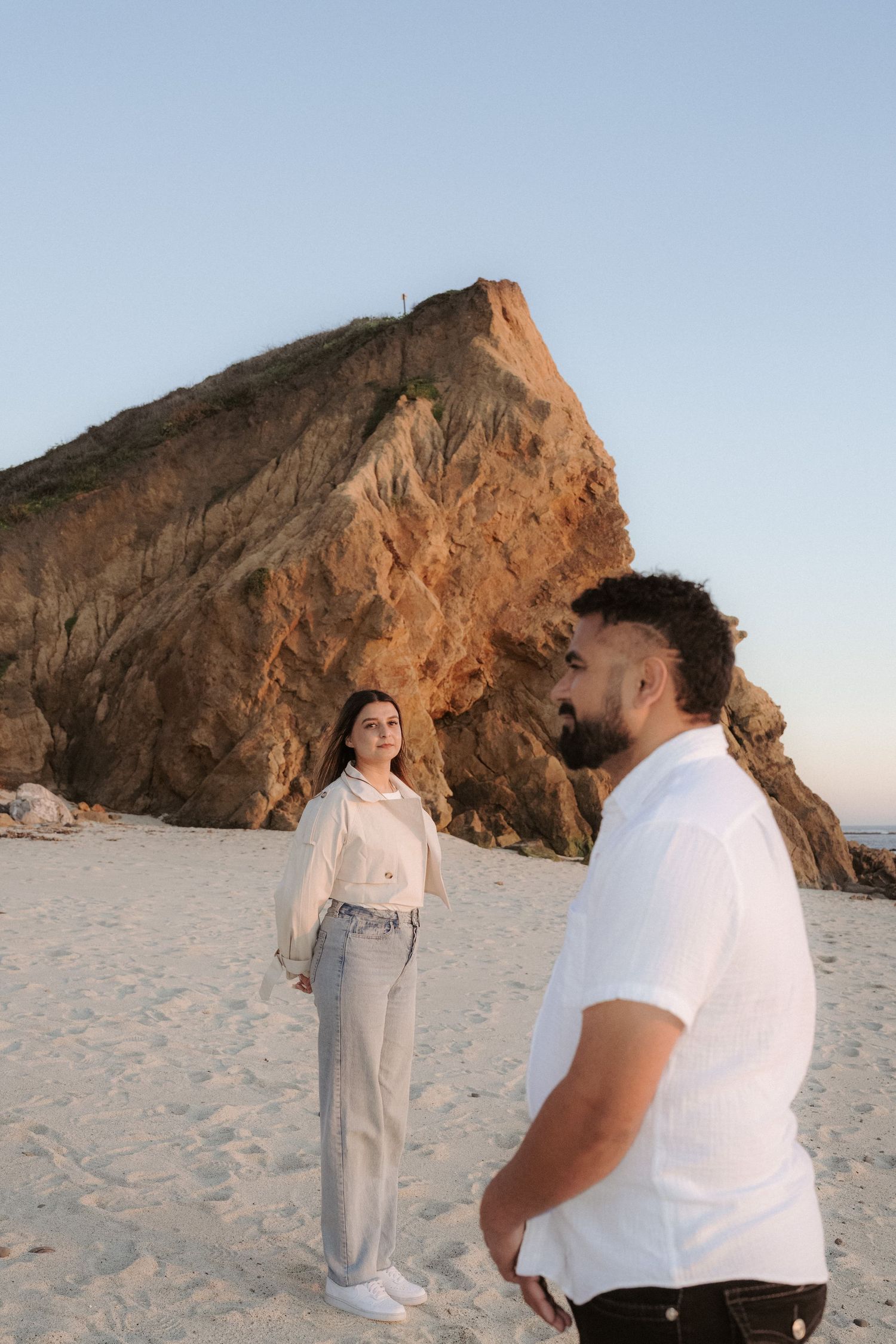 Couple standing on a sandy beach with a large rocky cliff backdrop at dusk, dressed in casual white outfits.