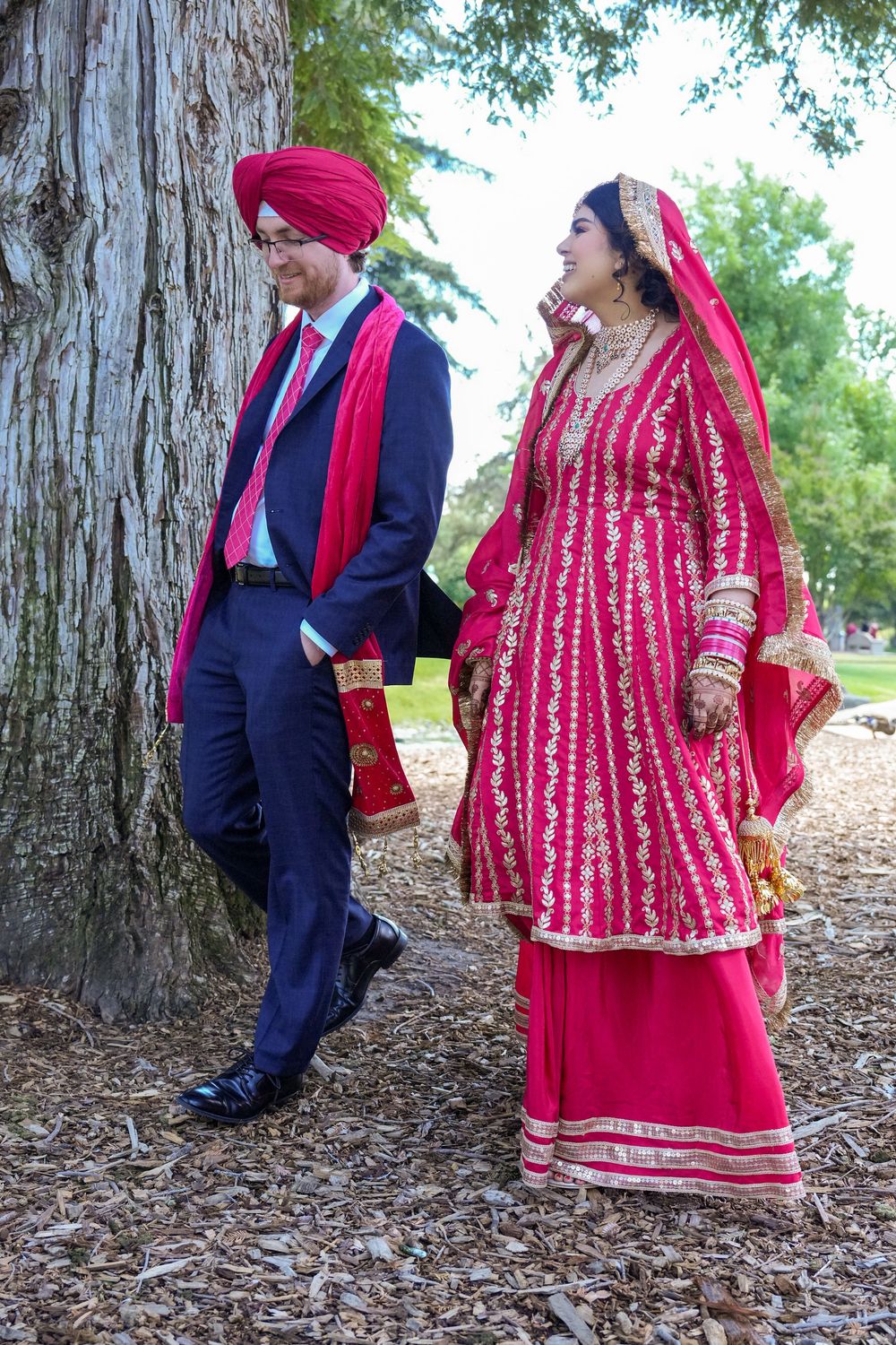 A couple in traditional Punjabi wedding attire stand near a tree trunk, with the bride wearing an elegant red lehenga.