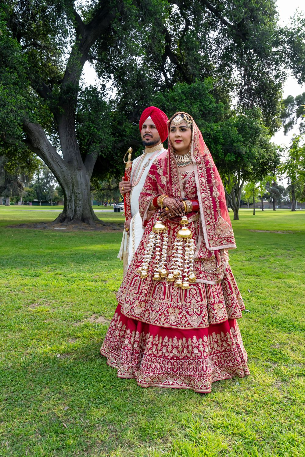 Traditional Sikh couple in wedding attire standing in a park with a majestic tree behind them.