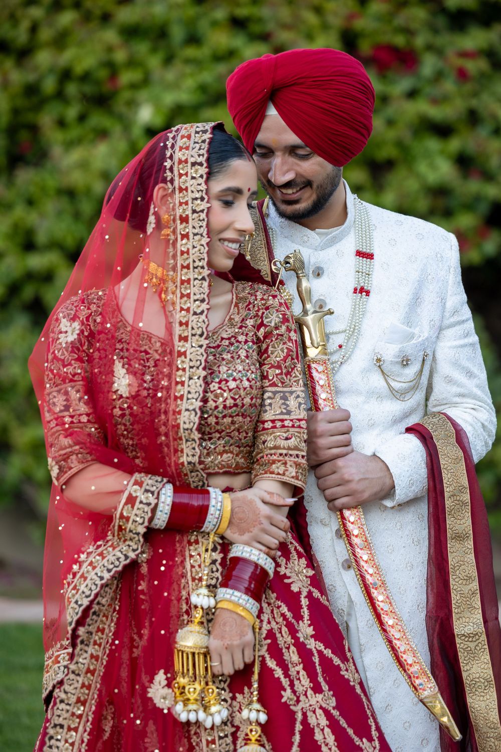 A couple in traditional Indian wedding attire standing together outdoors, bride in red lehenga and groom in white sherwani.