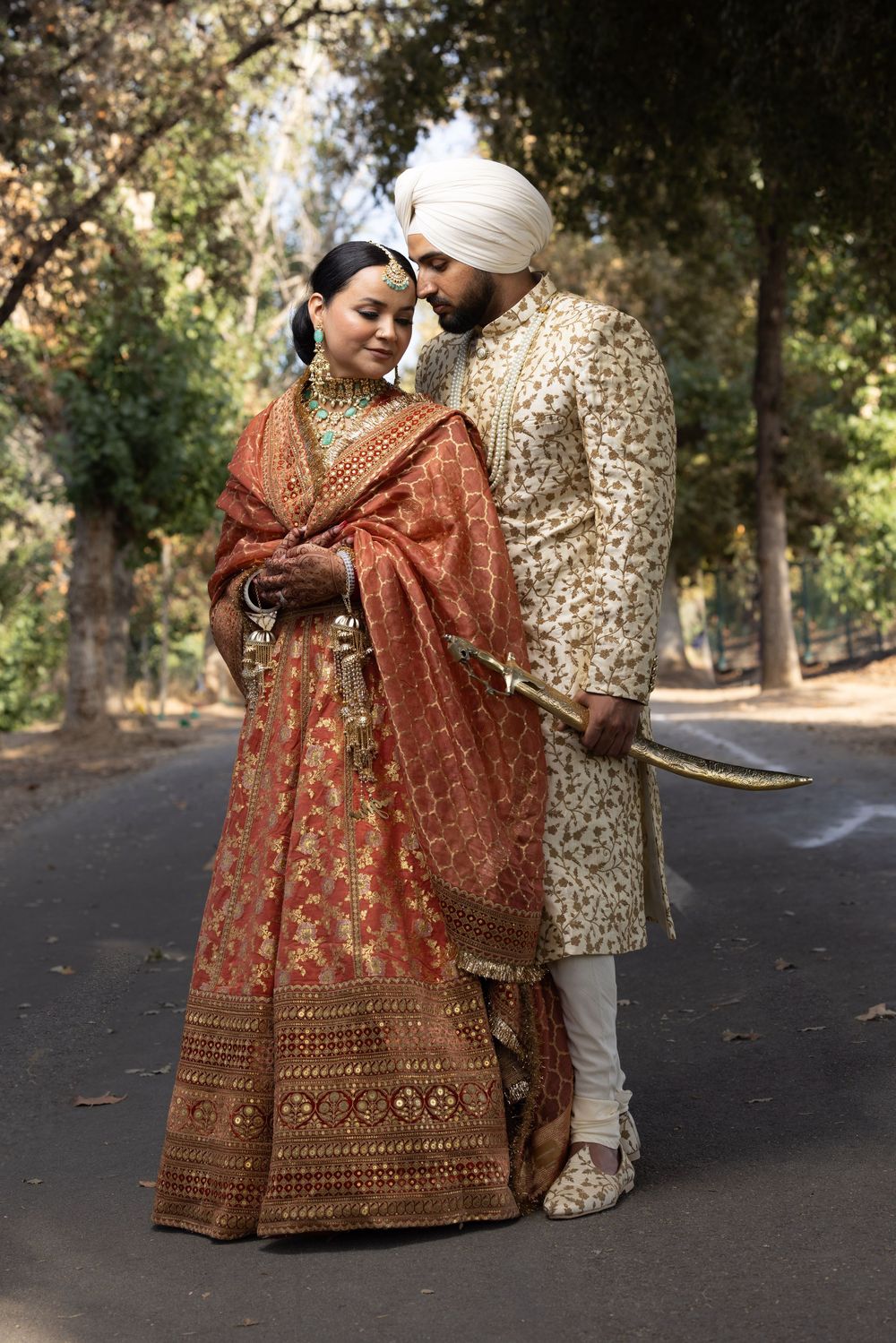A couple in traditional Indian wedding attire share an intimate moment on a tree-lined path during their wedding photoshoot.