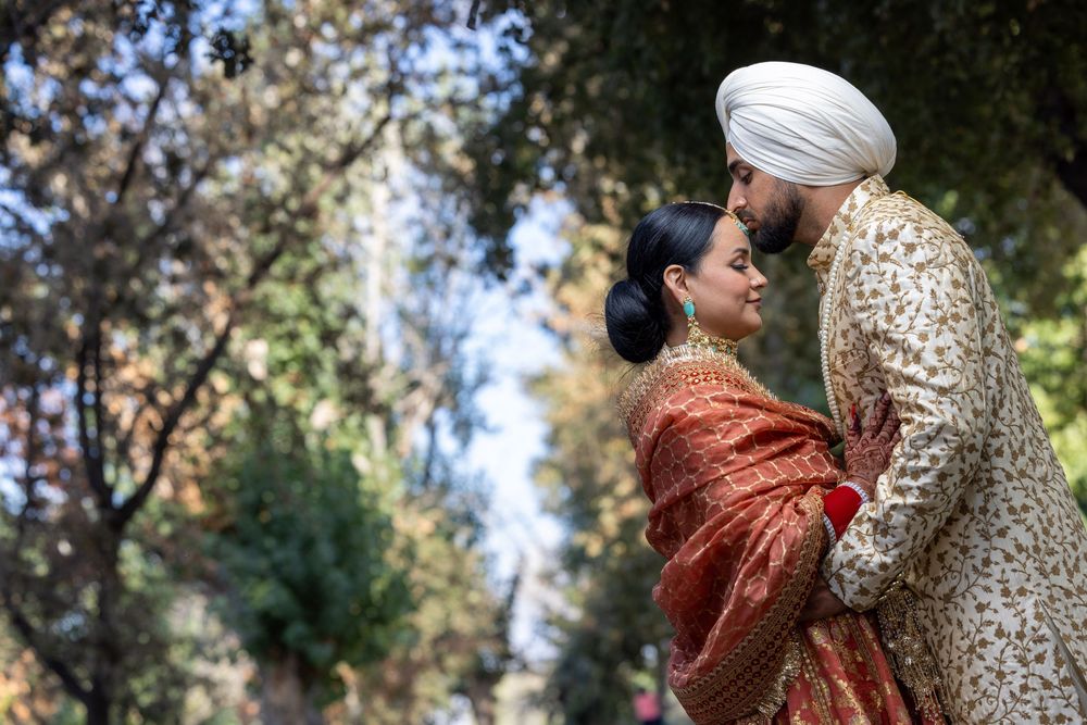 A couple in traditional South Asian wedding attire embrace outdoors surrounded by trees.