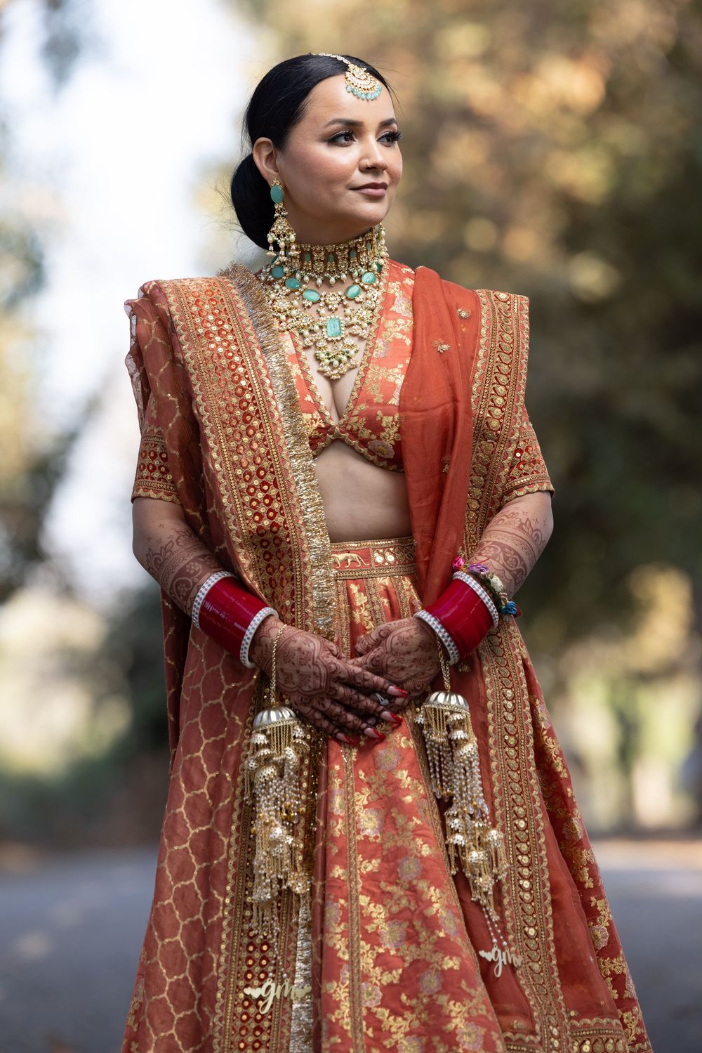 A bride in an ornate coral lehenga with intricate gold embroidery and traditional Indian bridal jewelry poses outdoors.