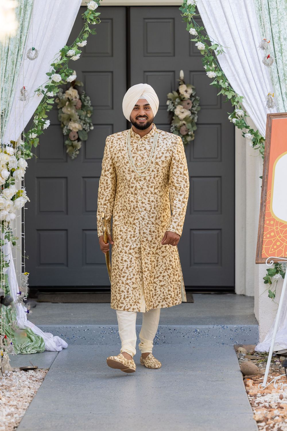 Traditional Indian groom wearing an ornate gold embroidered sherwani stands in front of a floral-decorated doorway.