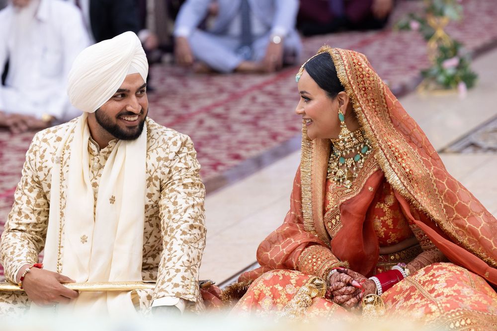 A couple in traditional Indian wedding attire, with the groom in gold sherwani and bride in coral red lehenga, seated together.