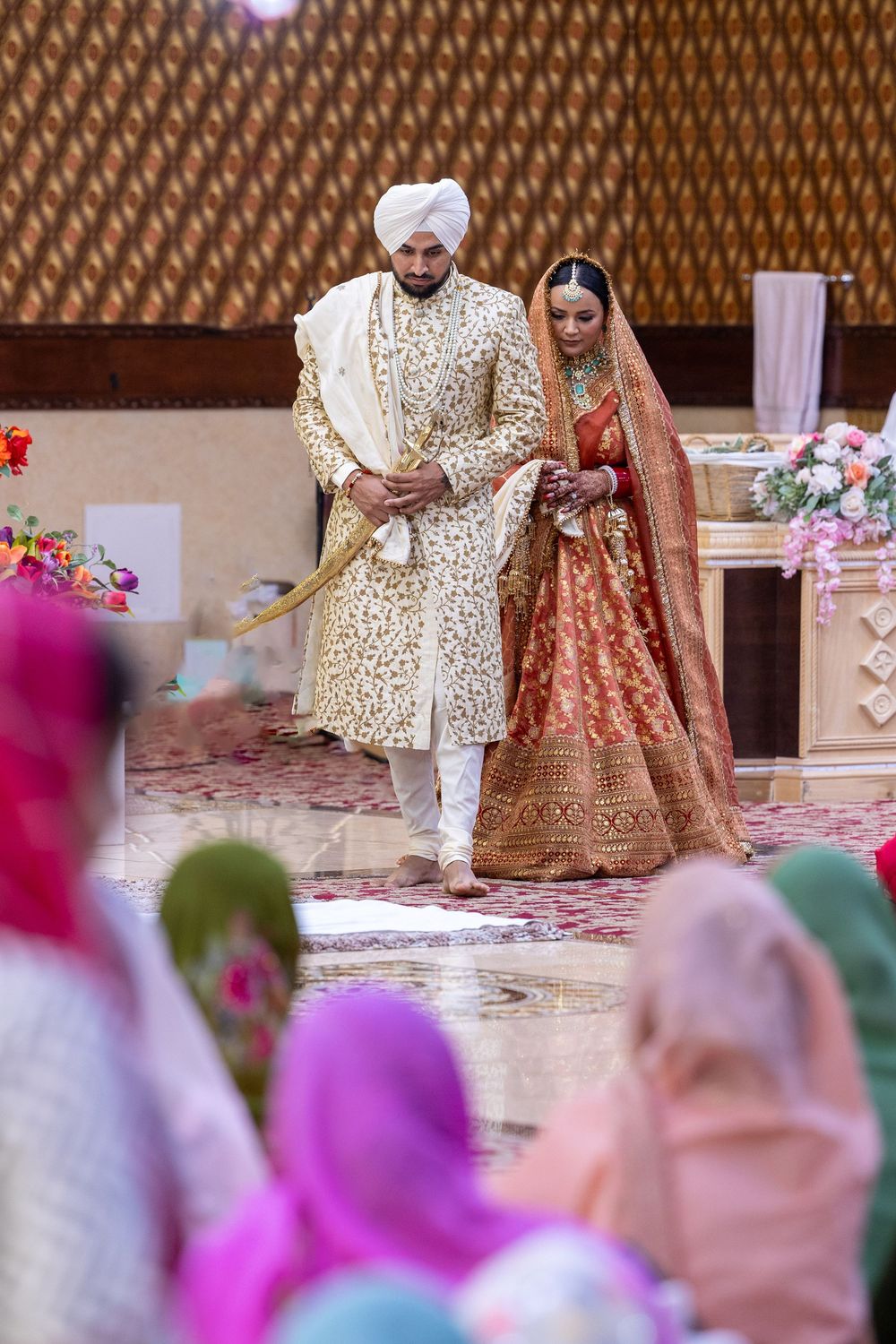 A couple in traditional Indian wedding attire walk together during a anand karaj ceremony as guests in colorful clothing look on.