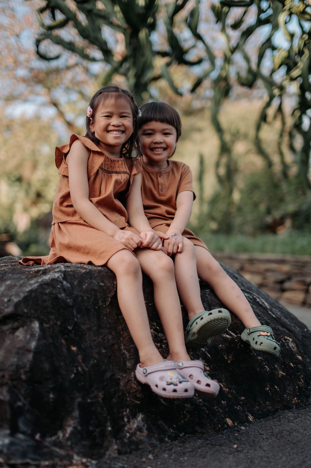 Two children in matching brown dresses sitting together and laughing on a large rock.