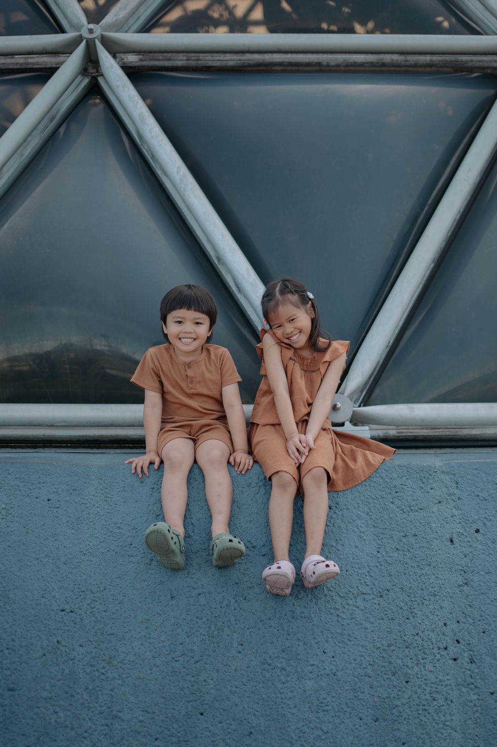 Two children in orange outfits sit together against a geometric wall structure outdoors.