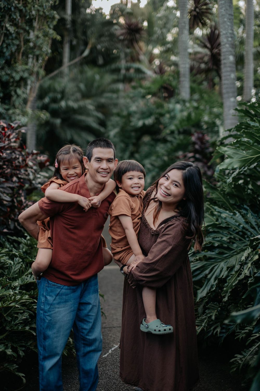A family stands together surrounded by lush tropical greenery in a garden setting during golden hour.