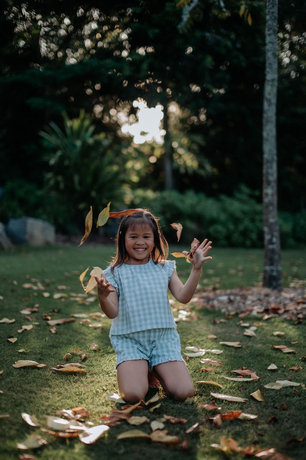 Young child in checkered outfit playing and waving while kneeling on fallen leaves.