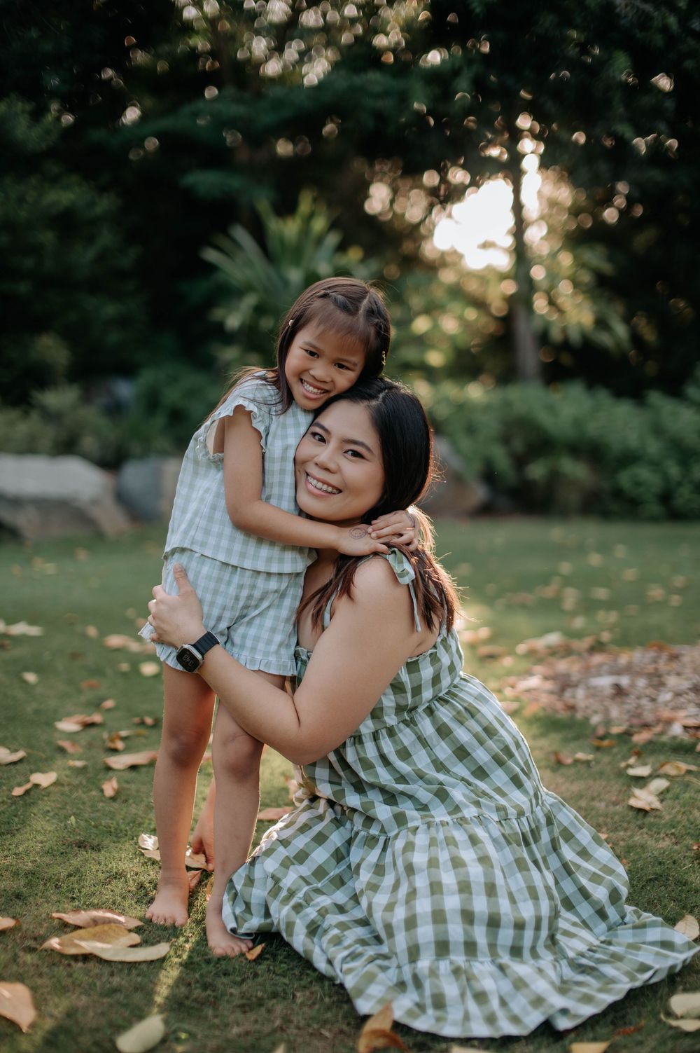 Two people in matching gingham dresses share tender moment outdoors during golden hour portrait session.