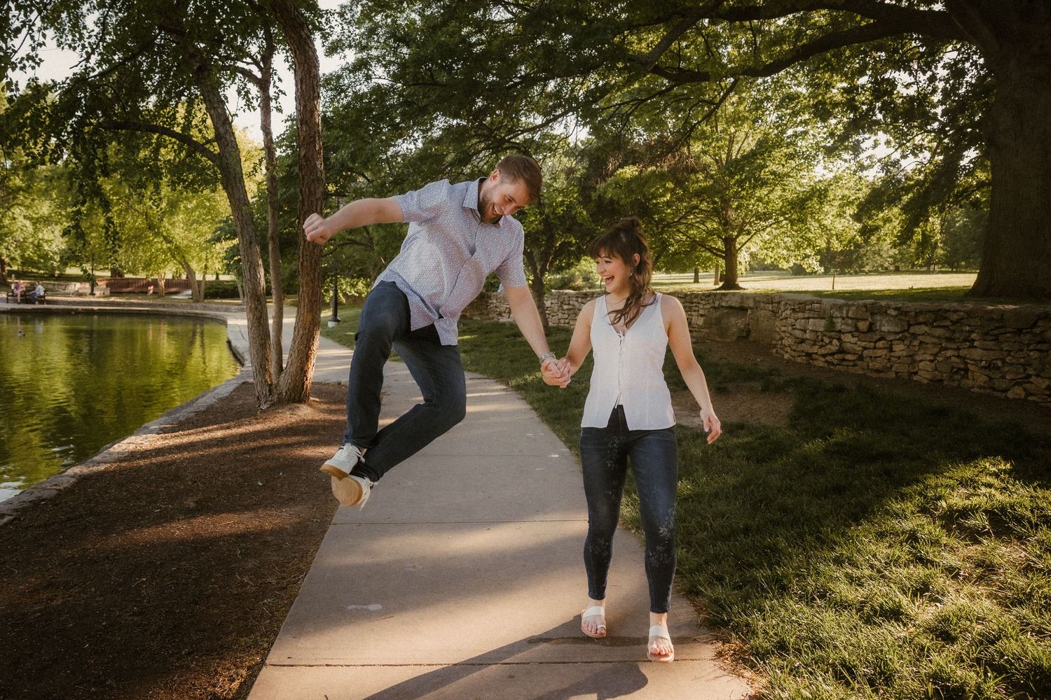 Two people playfully jumping together on a walking path beside a pond in a sunny park setting during golden hour. Shawnee Kansas photos.