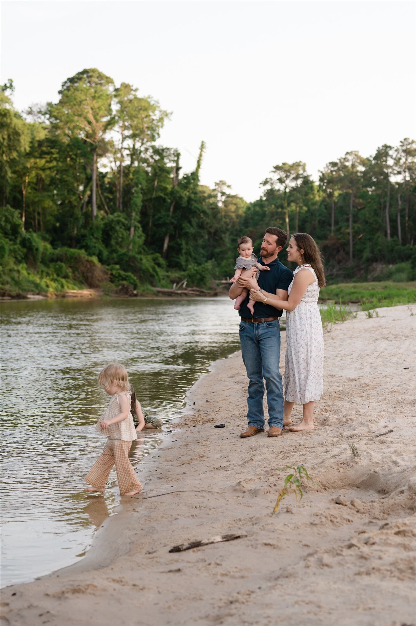 A family enjoys a peaceful evening stroll along a sandy riverbank surrounded by lush green forest.