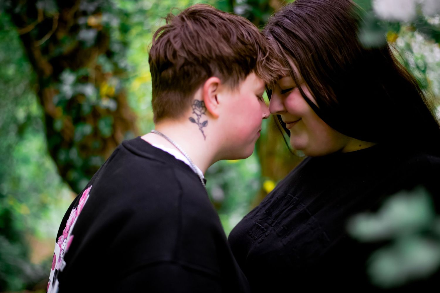 Two people share an intimate moment touching foreheads while wearing black clothing in a garden setting.