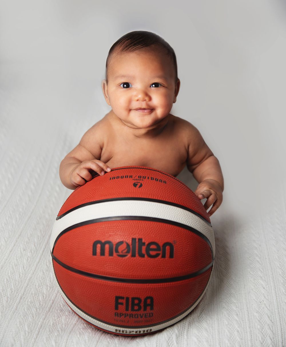 Happy baby sits with a red and white Molten FIBA regulation basketball on a light grey background. During a newborn photoshoot in Sidcup