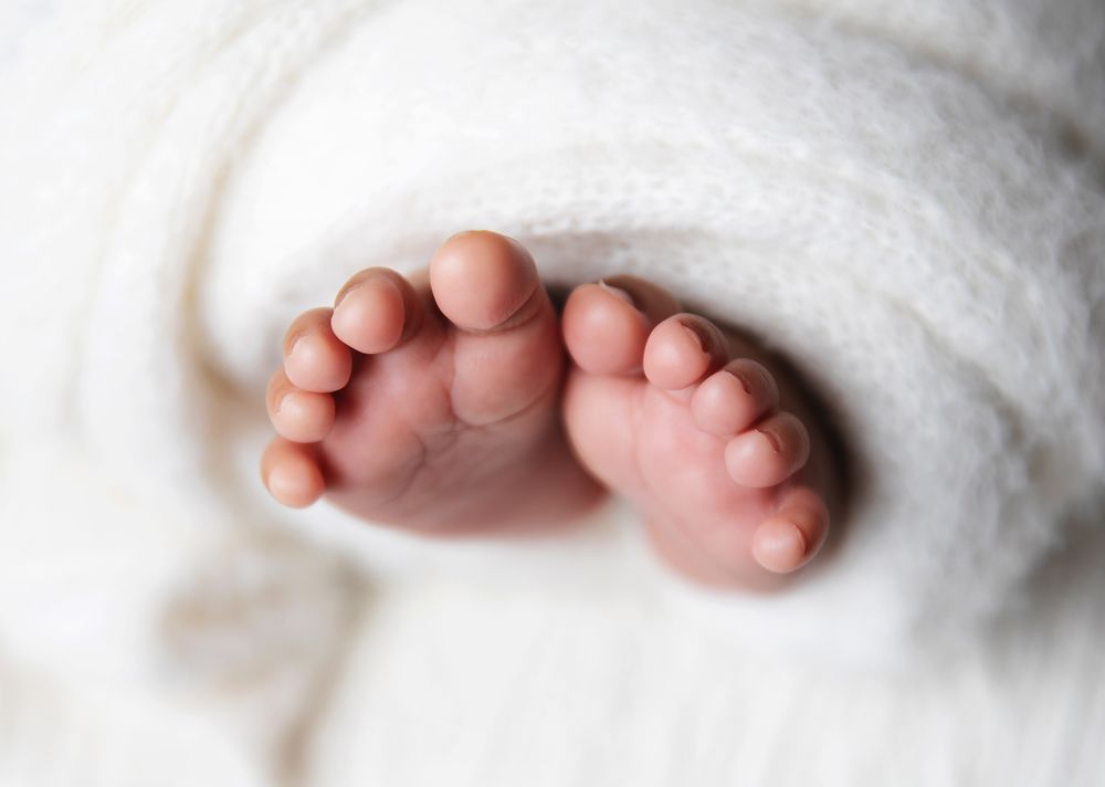 Tiny baby feet peek out from beneath a soft white blanket, showing delicate pink toes against a light background.