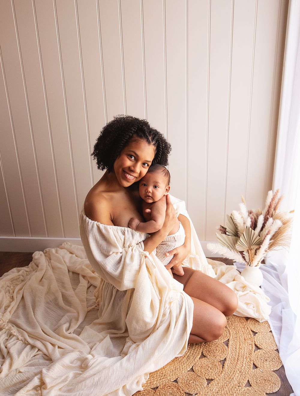 Mother and baby in matching white outfits sitting on a woven rug with pampas grass decor. During a newborn photoshoot in London